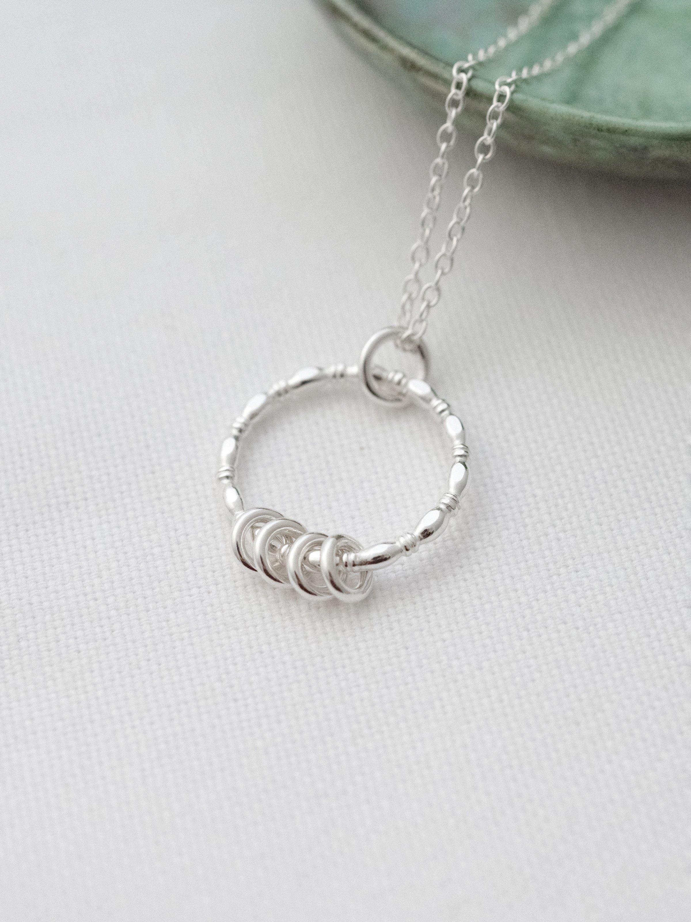 A close up view of a silver 4 ring necklace displayed on white material and a turquoise jewellery dish in the background.