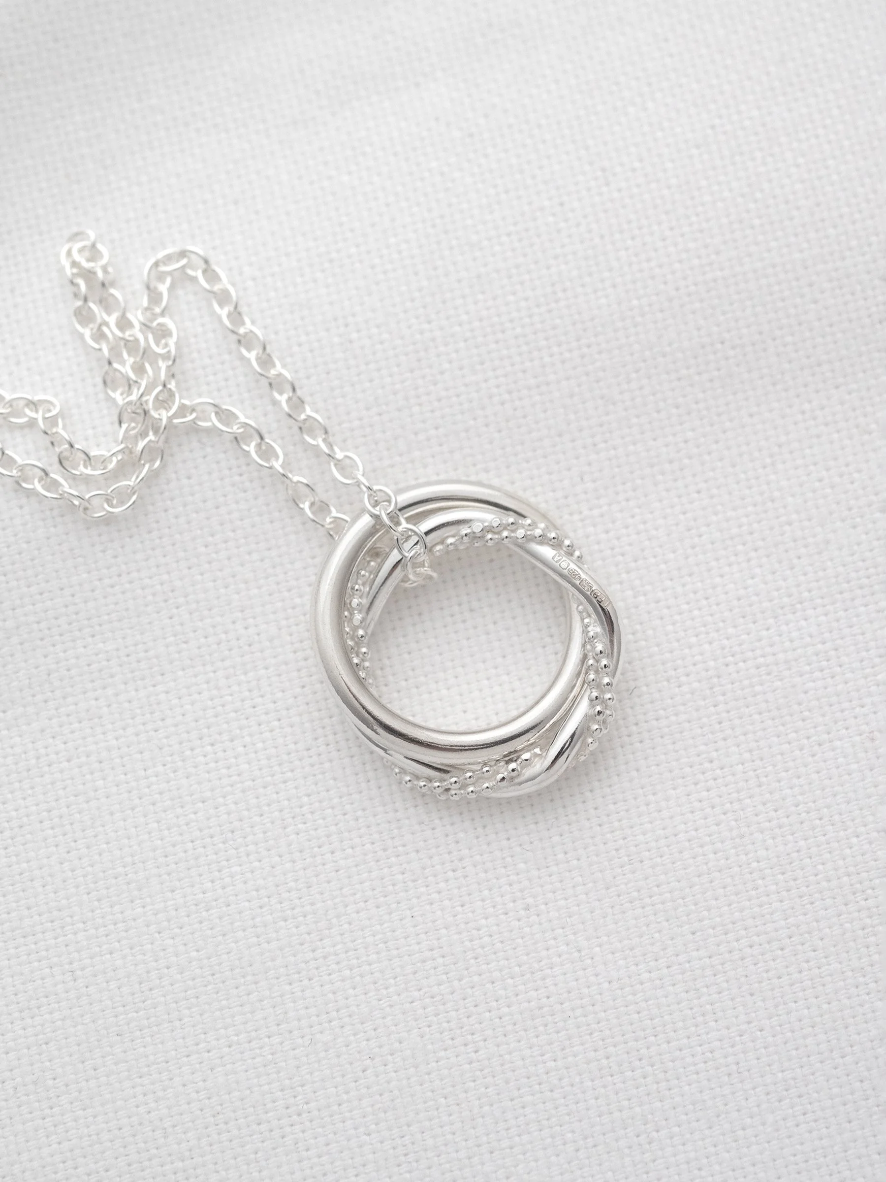 A close up of a silver interlinked two ring necklace displayed on white material.