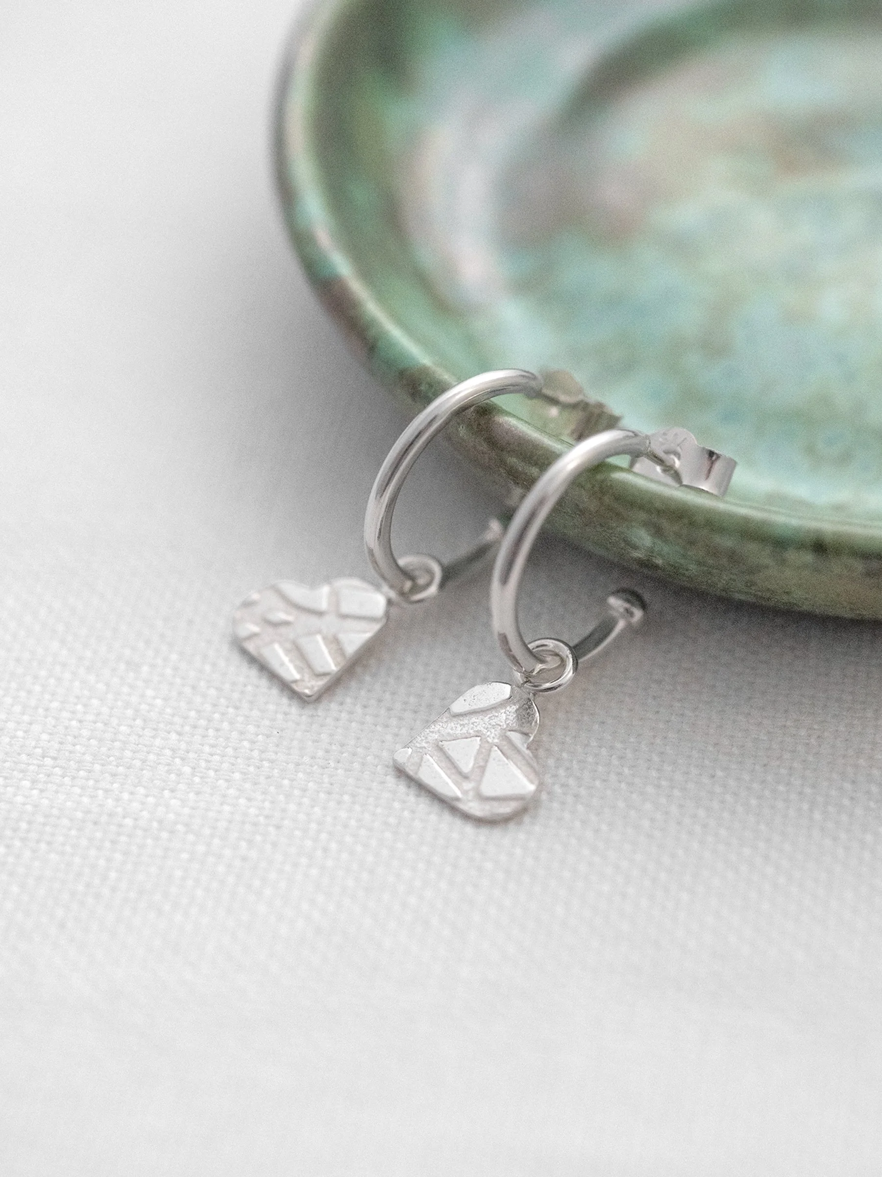A side view of silver heart hoop earrings hanging over a green jewellery dish.