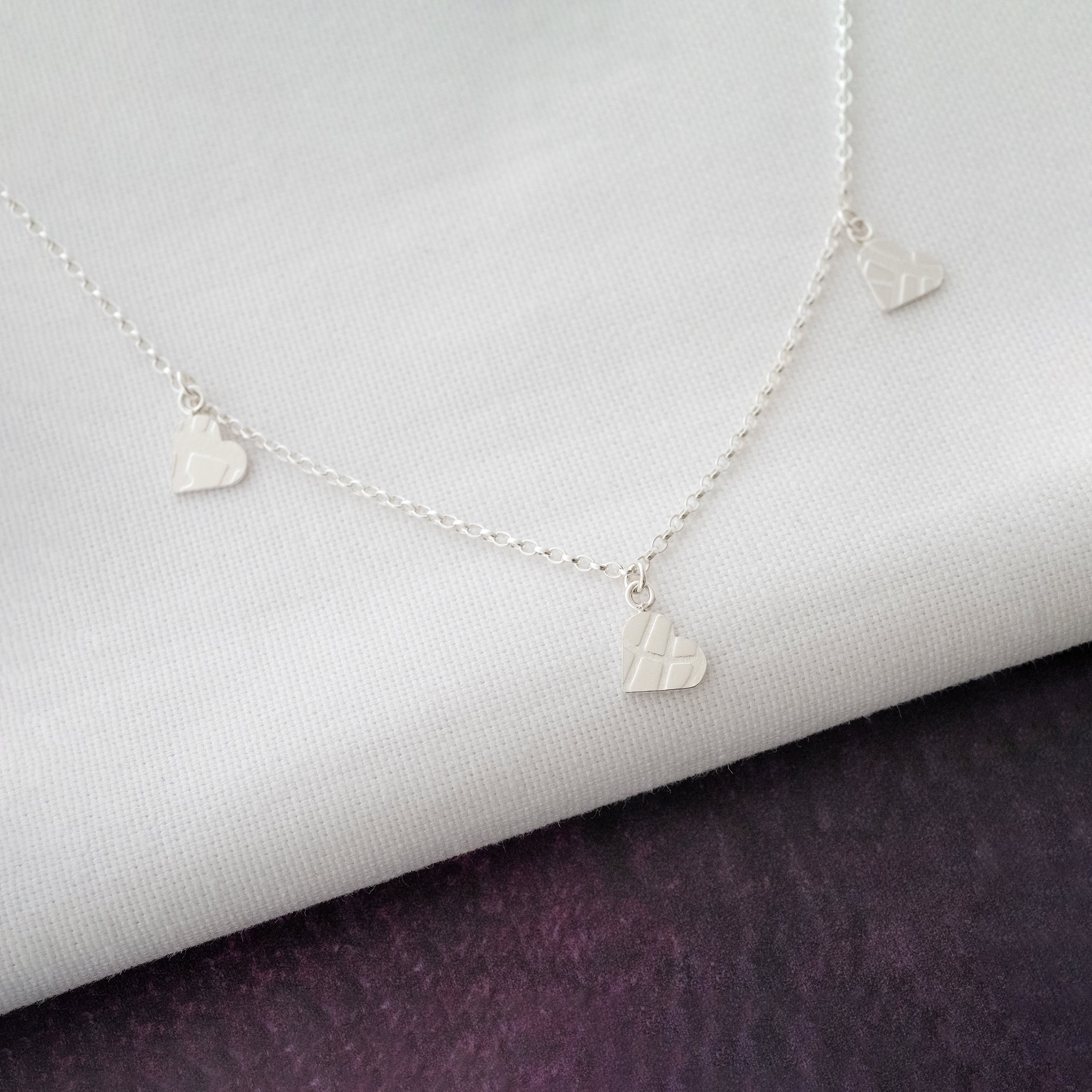A sterling silver three heart textured necklace displayed over white material and a purple background.