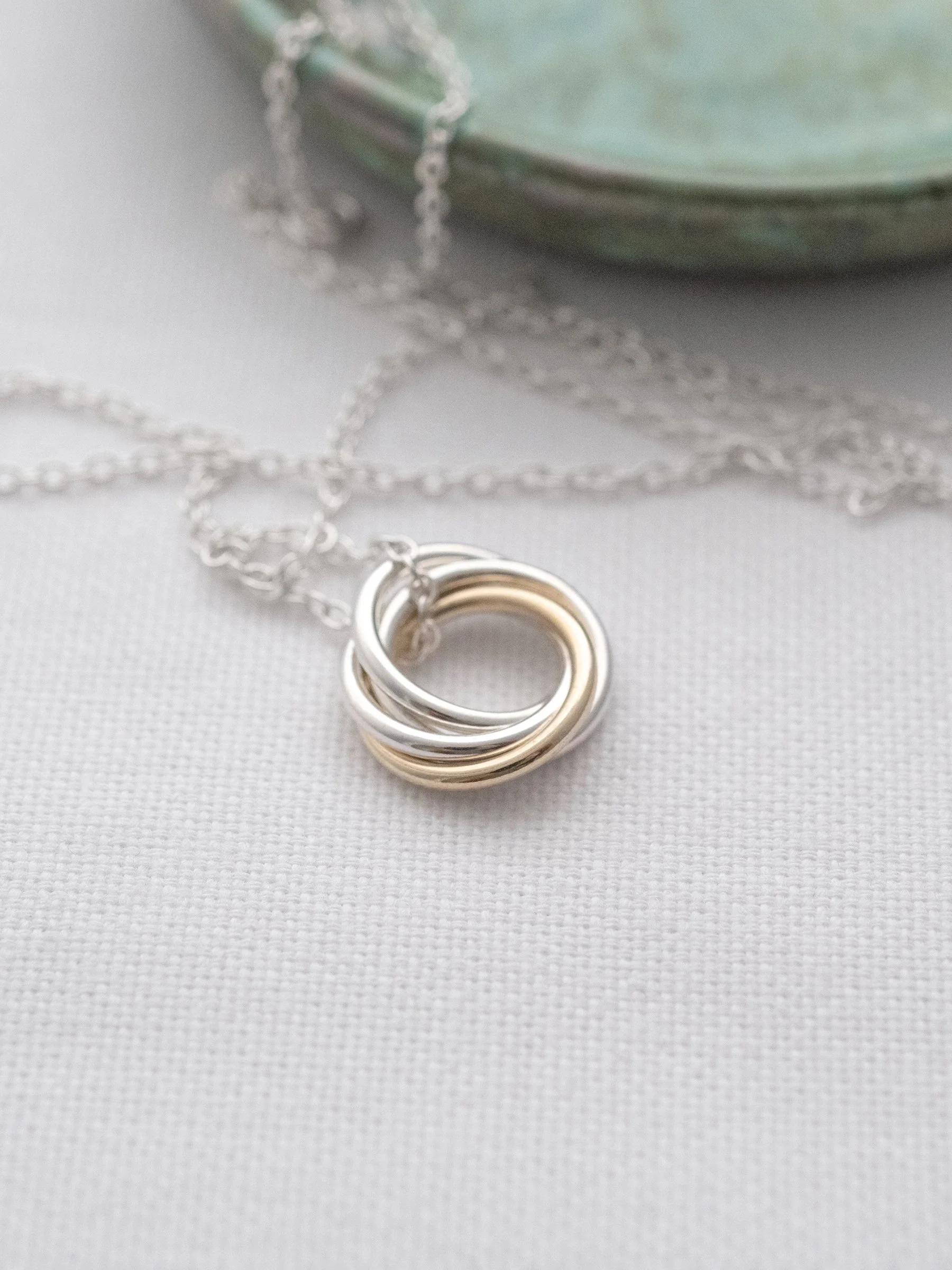 A gold and silver four ring necklace displayed on white material with a green jewellery dish in the background.