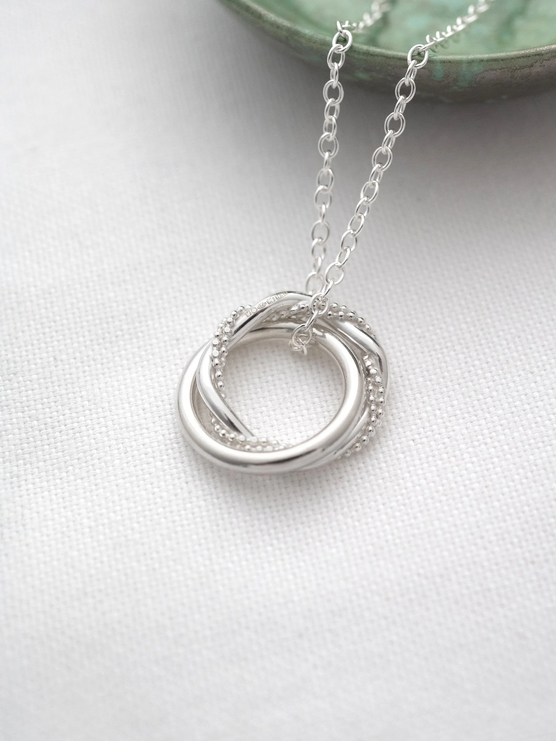 A close up of a silver interlinked necklace displayed on white material with a green jewellery dish in the background.