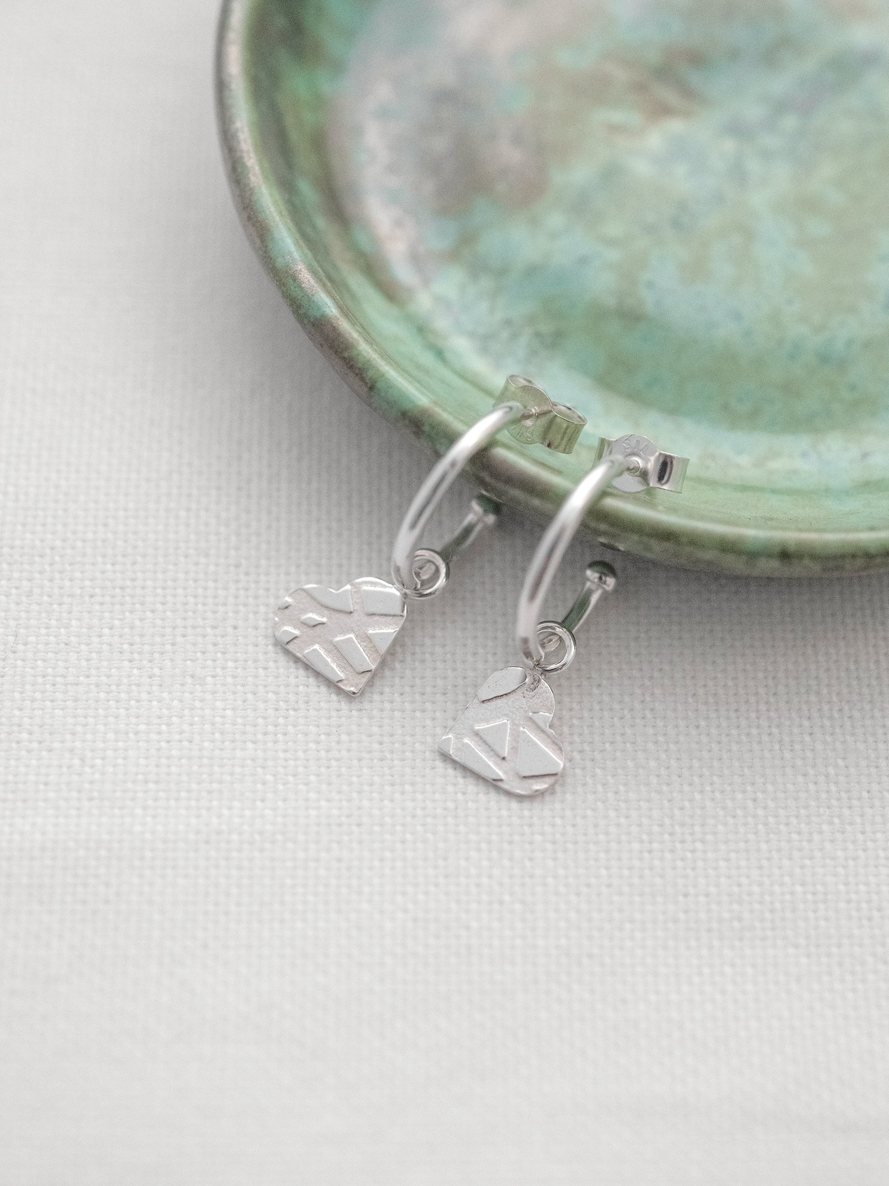A pair of silver patterned heart hoop earrings displayed over a small green jewellery dish.