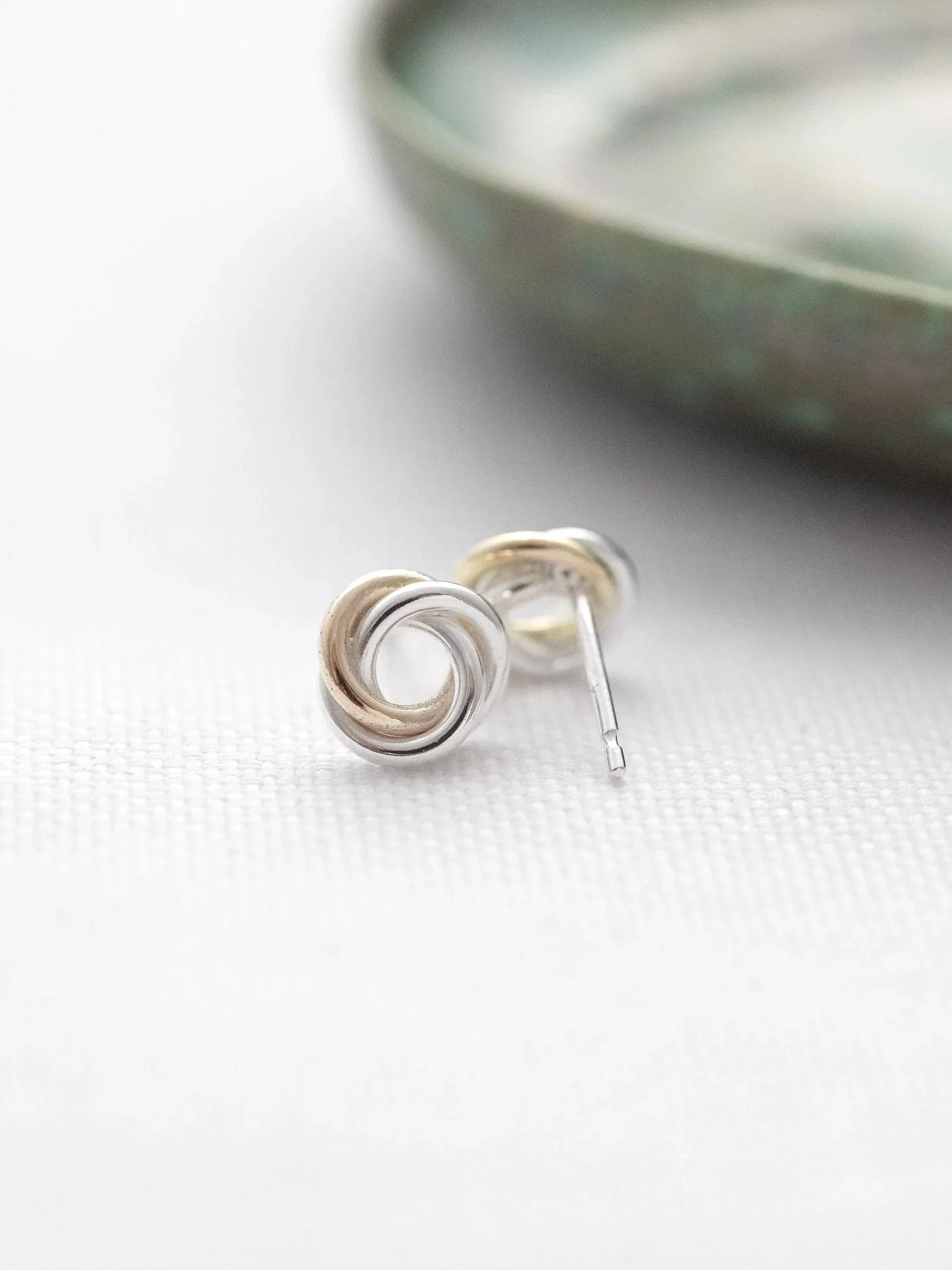 A front a back view of a pair of handmade gold and silver interlinked stud earrings displayed on white material with a green jewellery dish in the background.