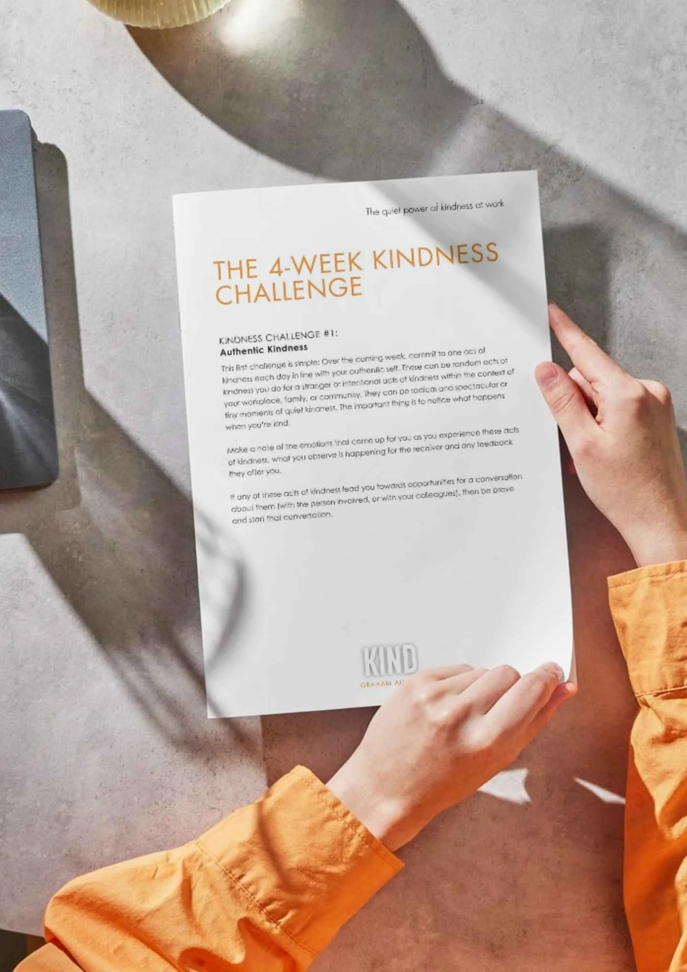 Person holding a sheet titled 'The 4-Week Kindness Challenge' on a desk with a laptop and a cup on the table.