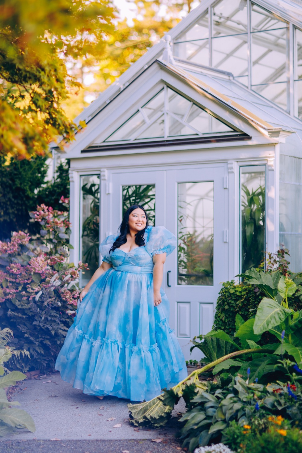 A plus-sized model in a Selkie dress posing in front of a greenhouse in direct sunlight, featuring a soft haze effect from a reflective prop in the corner.