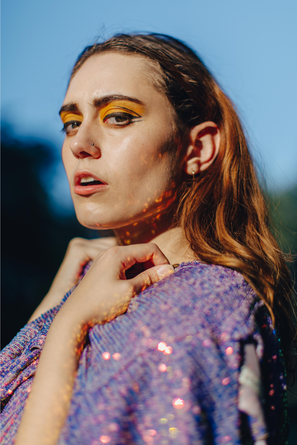 A close-up portrait of a model draped in sequined fabric to create an iridescent, mermaid-like shine and texture in harsh midday sun.
