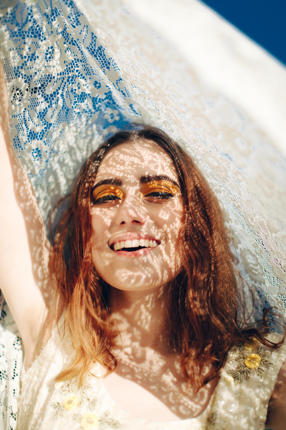 A model holding a thrifted vintage crochet tablecloth up to the sky to cast intricate lace patterns and shadows on her face in direct sunlight.