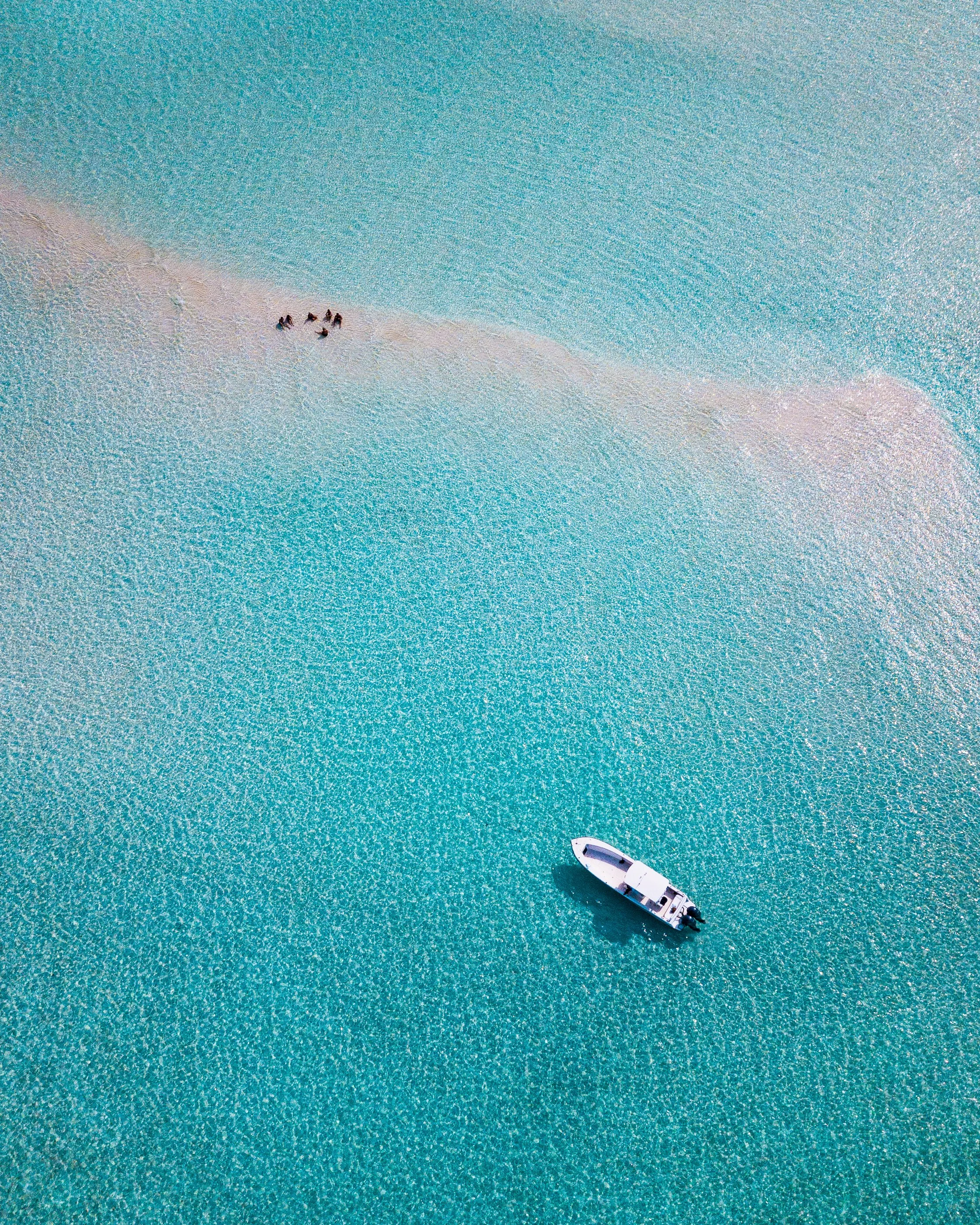 Aerial view of a boat floating on turquoise water with a small sandy island and four people on the beach in the distance.