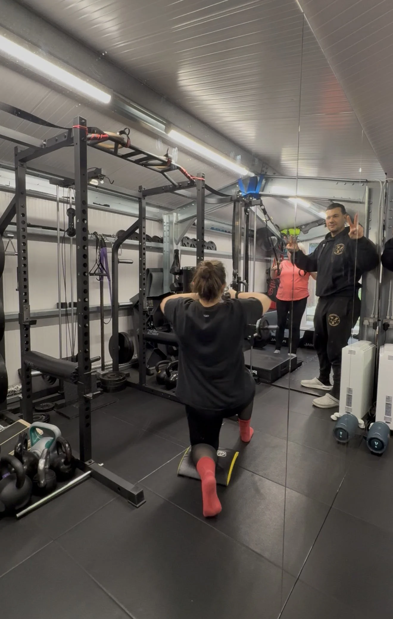 A woman is performing a squat exercise in a gym, facing a mirror. A man, possibly a trainer, is standing nearby, making a peace sign and smiling. The gym is equipped with various workout equipment and weights.