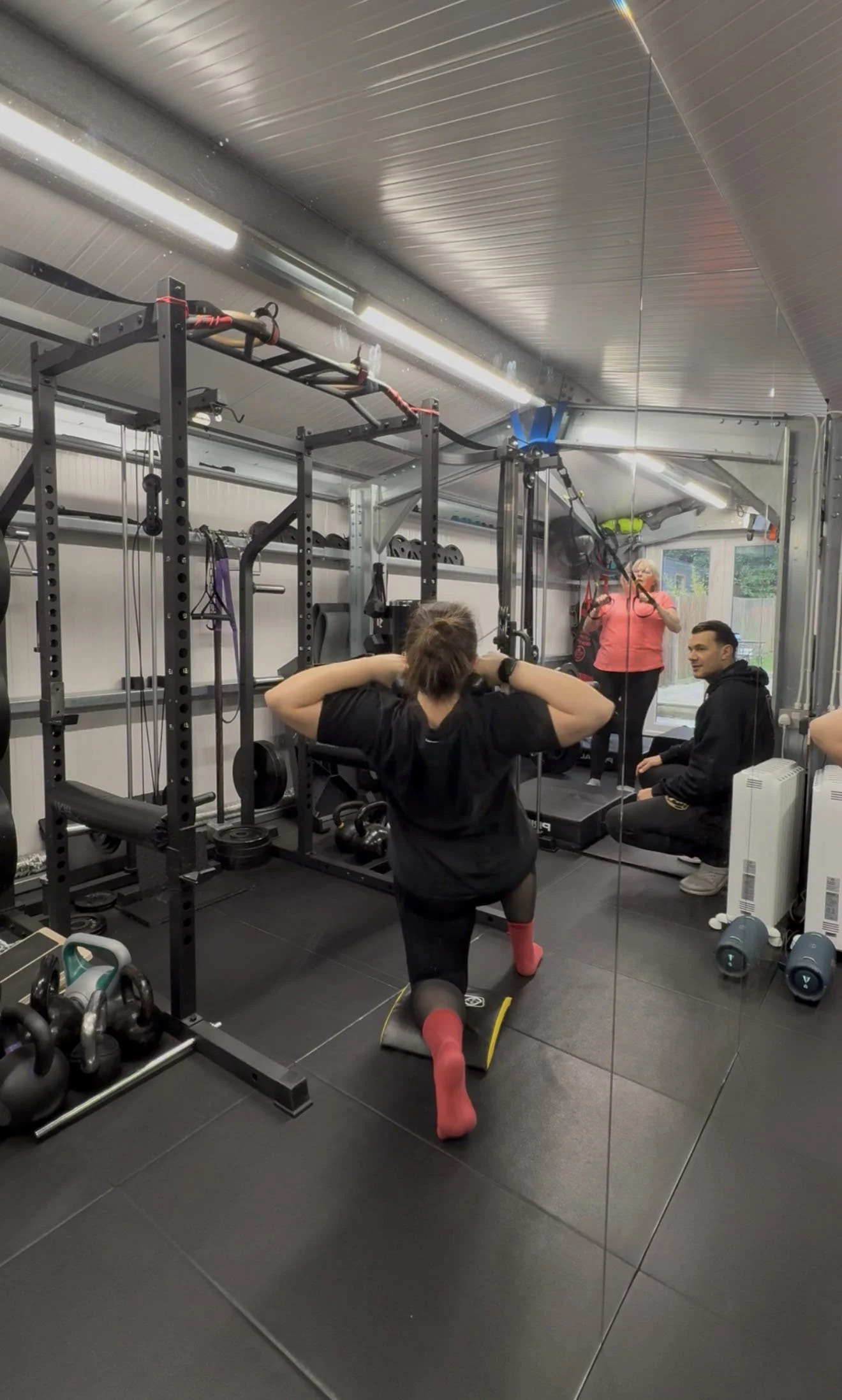 Woman doing hip exercises on a mat in a gym, with two women watching and a woman in the background using suspension straps.