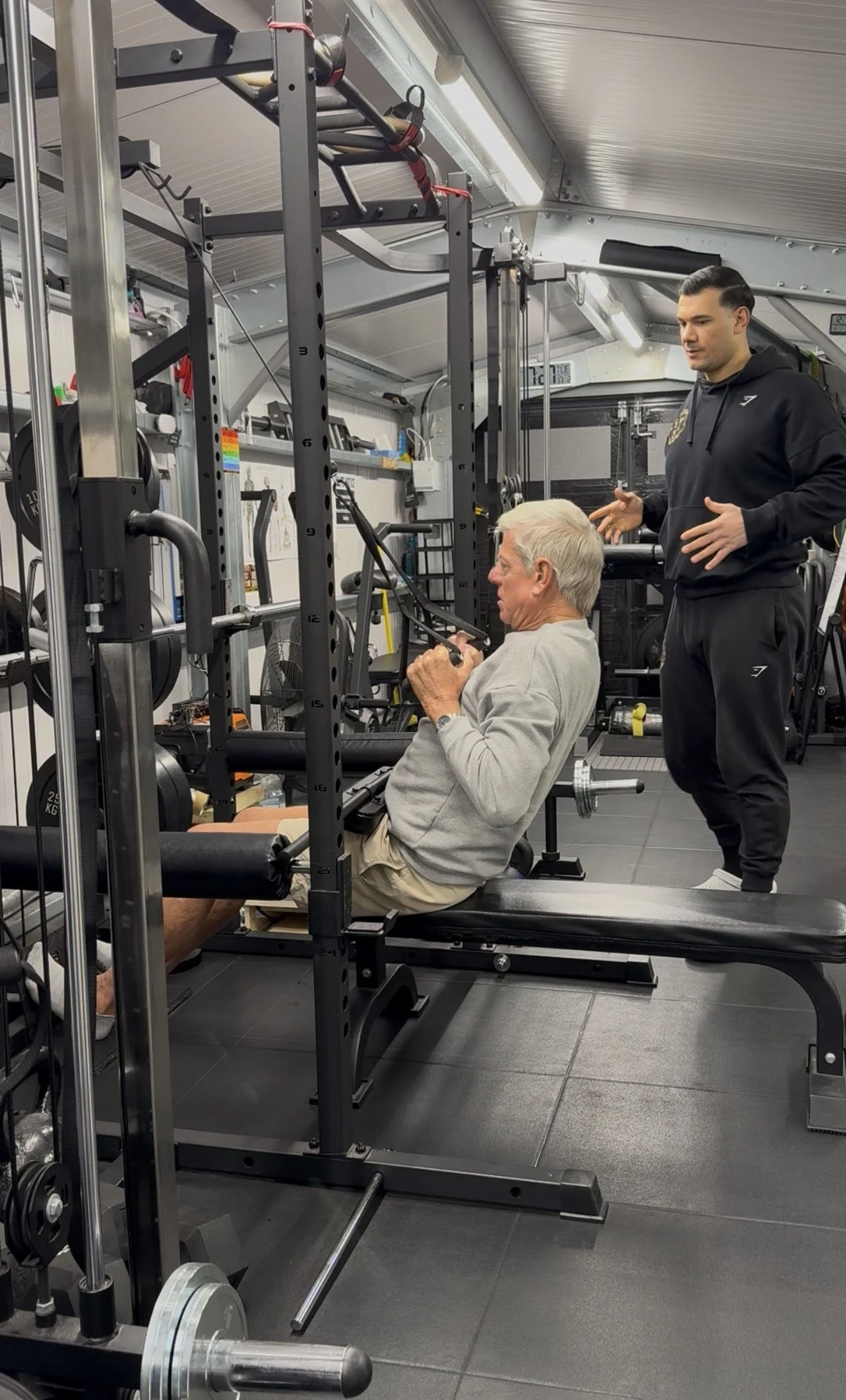 An elderly man working out on a fitness machine at a gym, with a personal trainer standing nearby, providing guidance. The gym is equipped with various weights and exercise equipment.