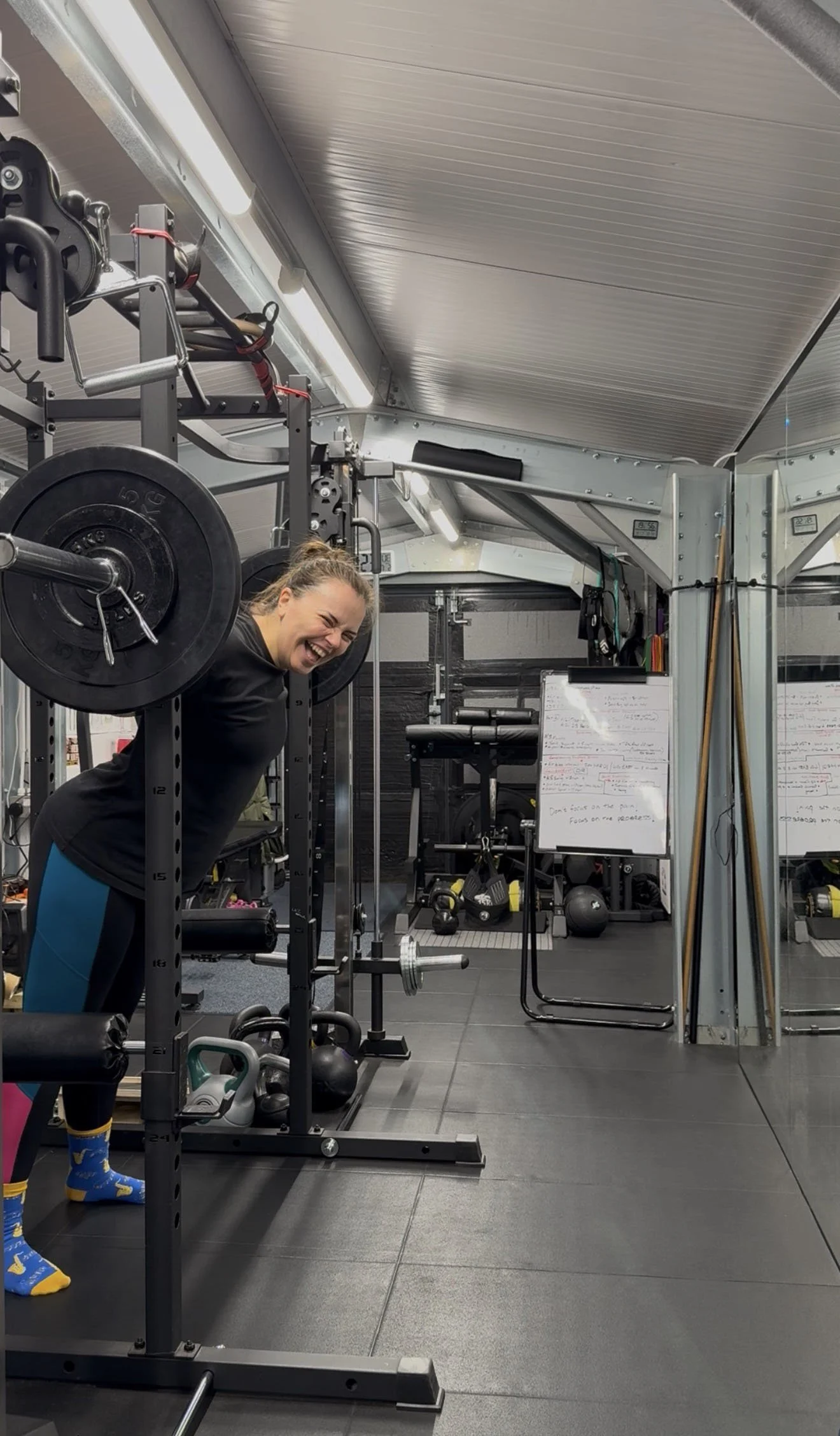 A woman in a gym lifting weights, smiling, and wearing colorful socks, with fitness equipment and a whiteboard in the background.