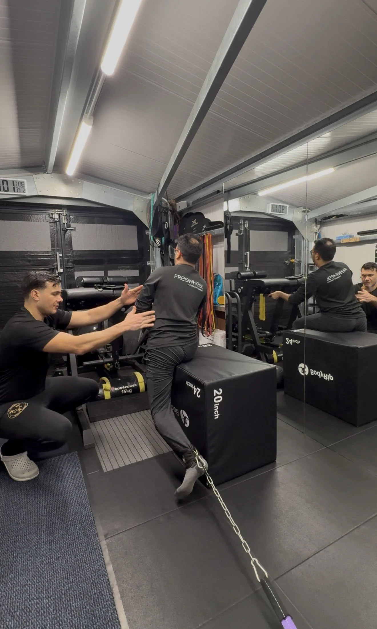 Two men are seated on boxes, pulling on weighted chains attached to their waist, while a trainer stands nearby giving instructions inside a gym. The gym has mirrors on the wall and various exercise equipment.