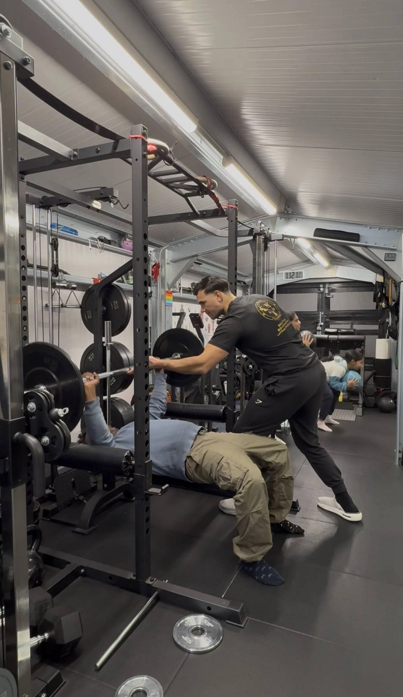 A person lying on a bench press at a gym lifting weights, with a person assisting them, and other people exercising in the background.