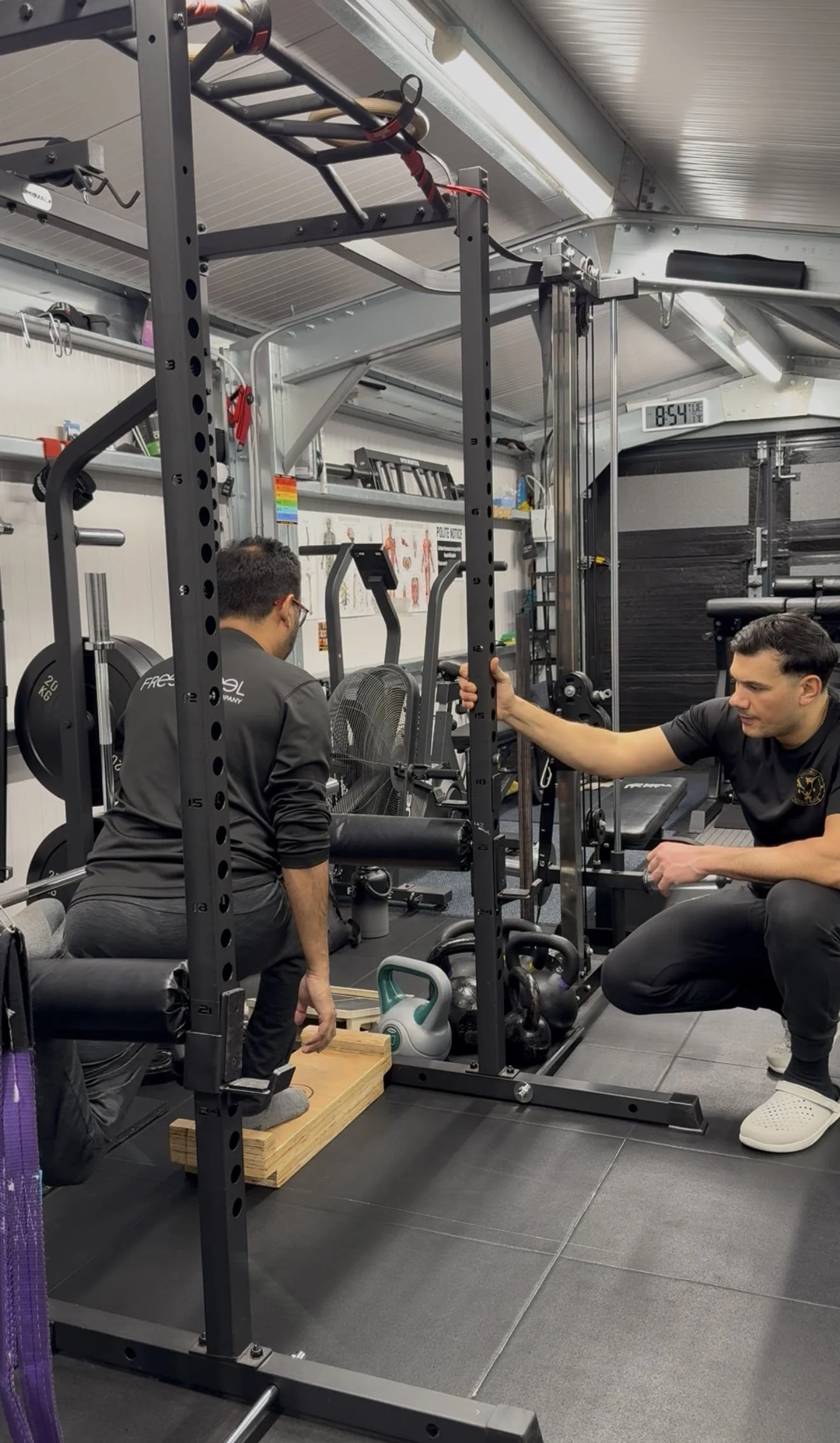 Two men in a gym training with a weightlifting machine. One man is kneeling on a wooden platform and the other is seated, holding onto the machine's bar. Gym equipment, kettlebells, and a fan are visible in the background.