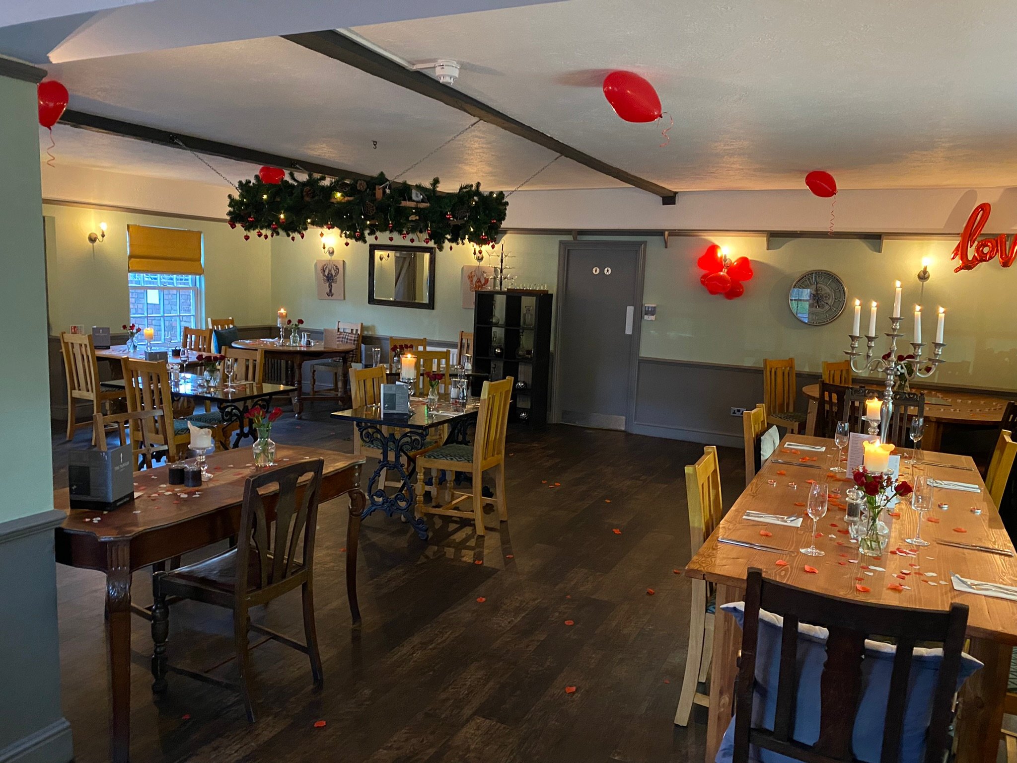 Romantic restaurant interior with wooden tables and chairs, decorated with red balloons and rose petals, dim lighting, and candlelit tables.