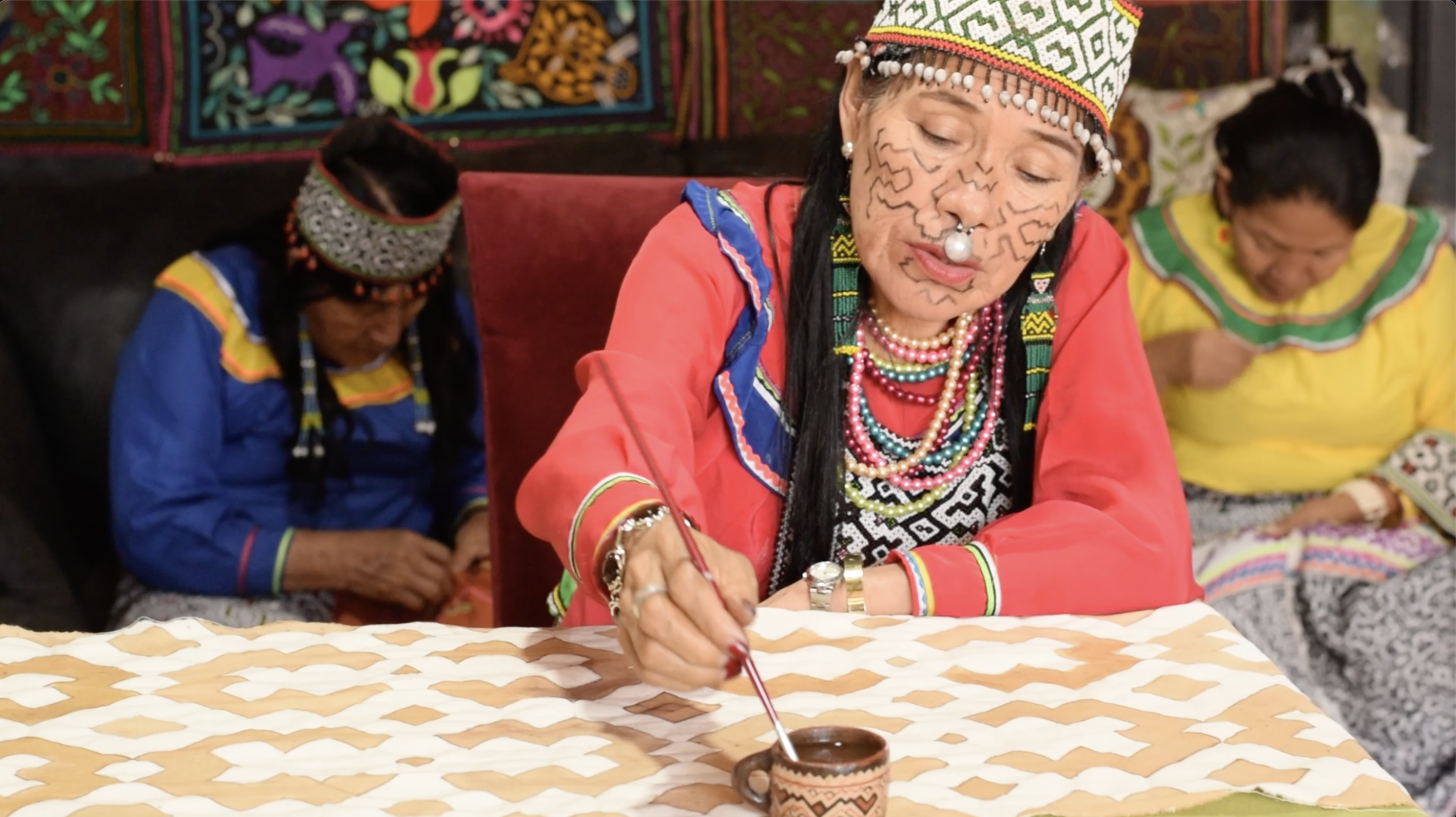 Women in traditional colorful attire, with face paint and jewelry, sitting at a table and engaging in painting or writing on a ceramic cup.