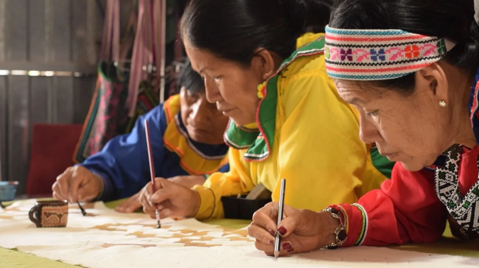 Three women dressed in traditional colorful clothing are focused on a craft project, writing or drawing on a large piece of fabric or paper, seated at a table in a rustic setting.