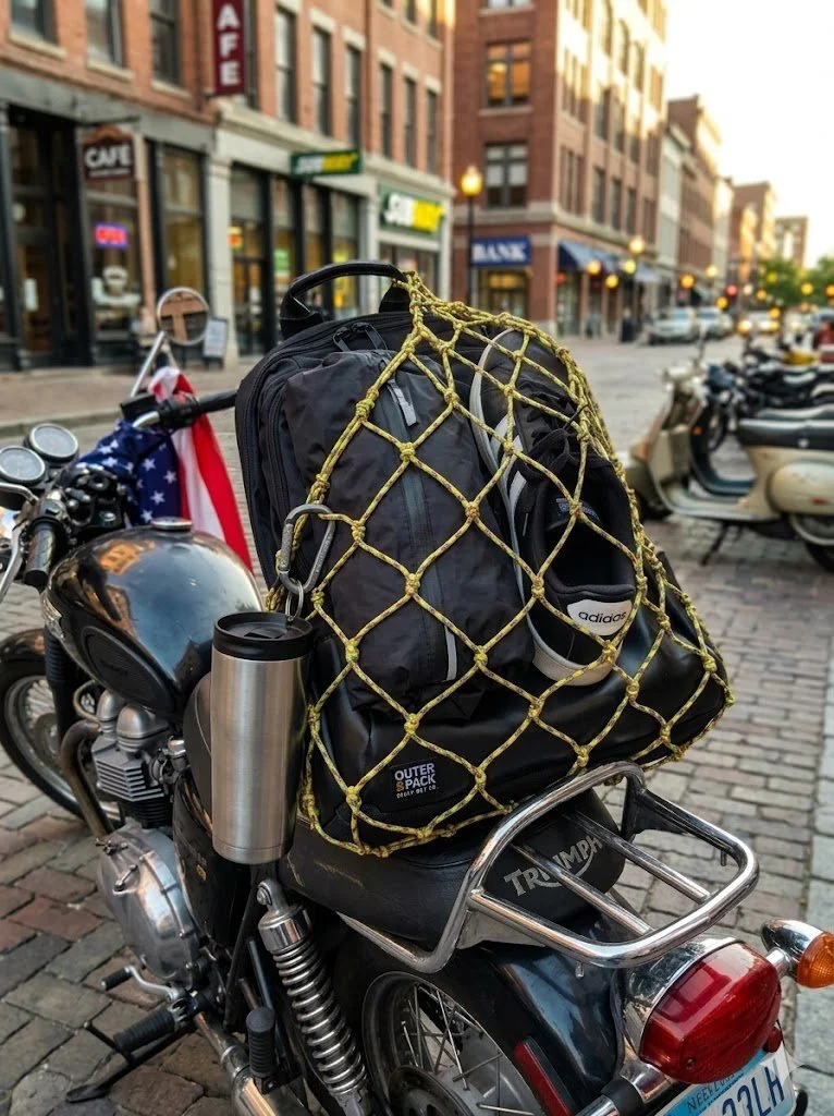 black backpack with Vietnam Vet color backpack net on a motorcycle with sneakers and a dry bag stuffed in it, and a water bottle clipped to one of the meshes.