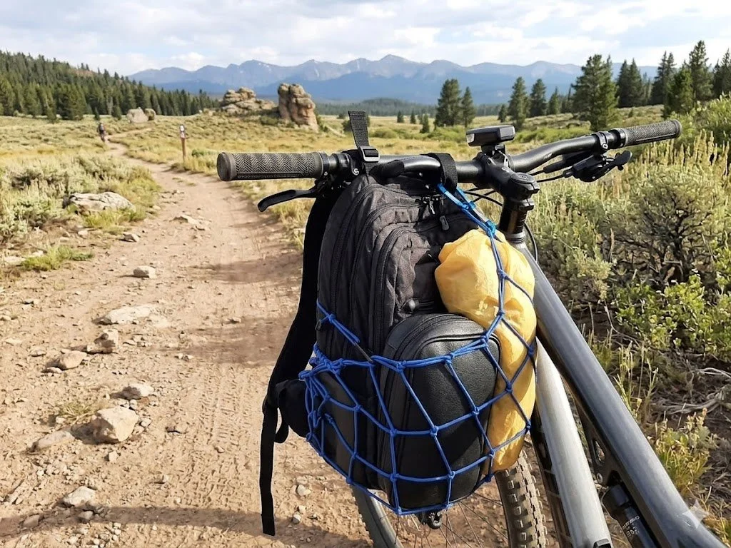 A black backpack with a blue backpack net holding a leather case and a yellow dry bag inside of it while hanging on the handlebars of a mountain bike