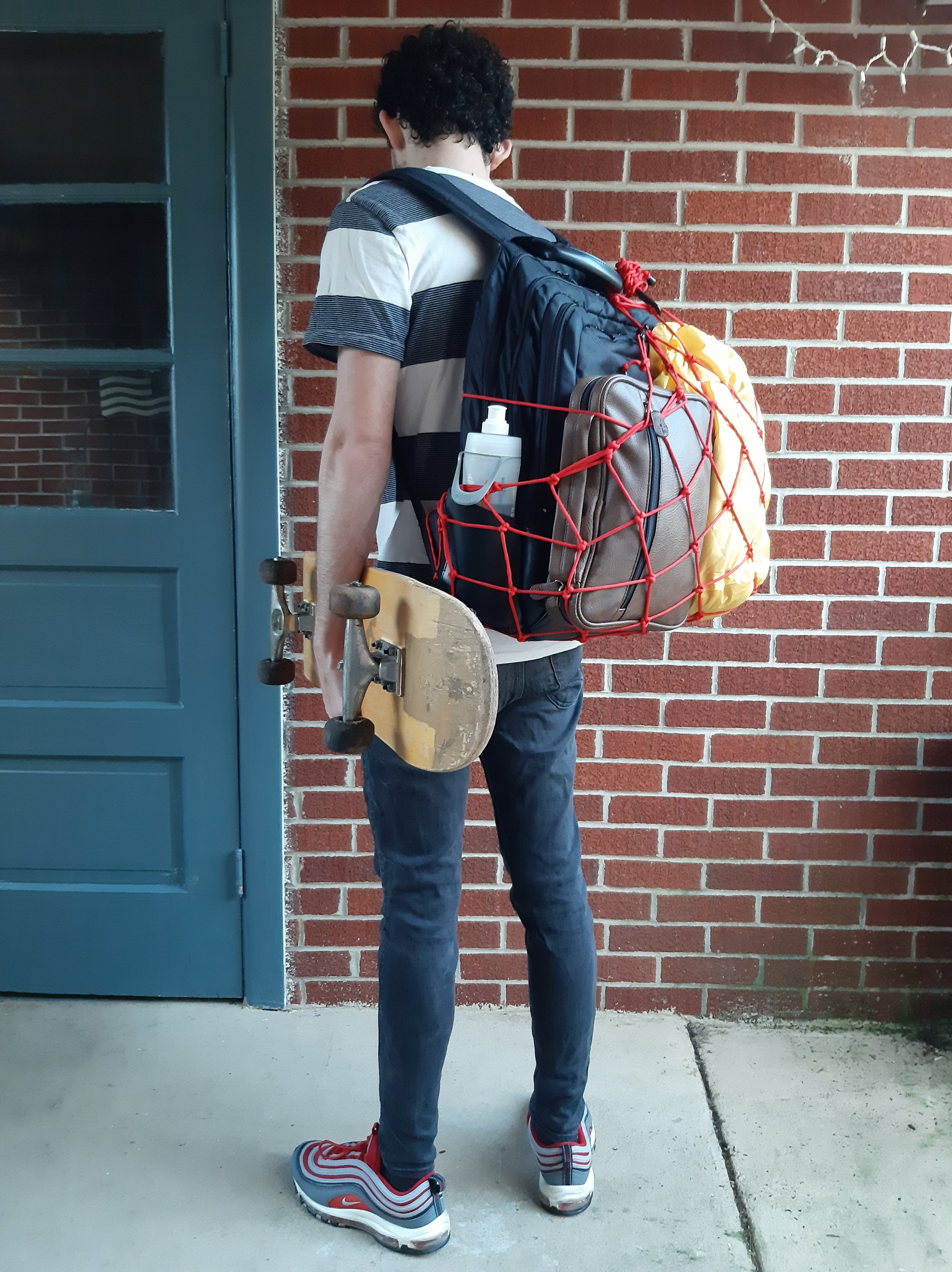 Person standing with backpack and skateboard next to brick wall and blue door.