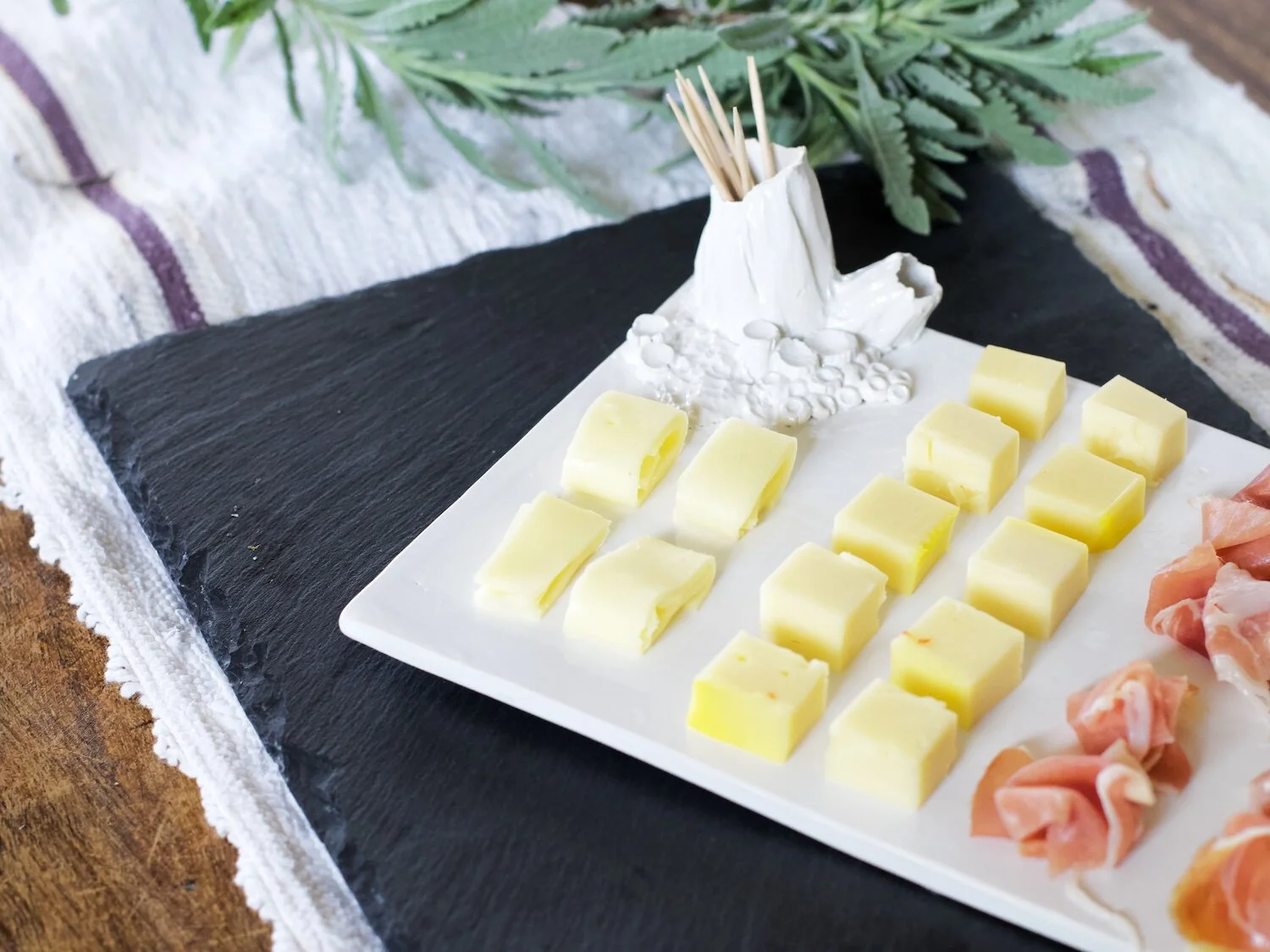 Prato de tapas branco com pormenor de recifes em cerâmica, em cima de mesa, com queijos e presunto