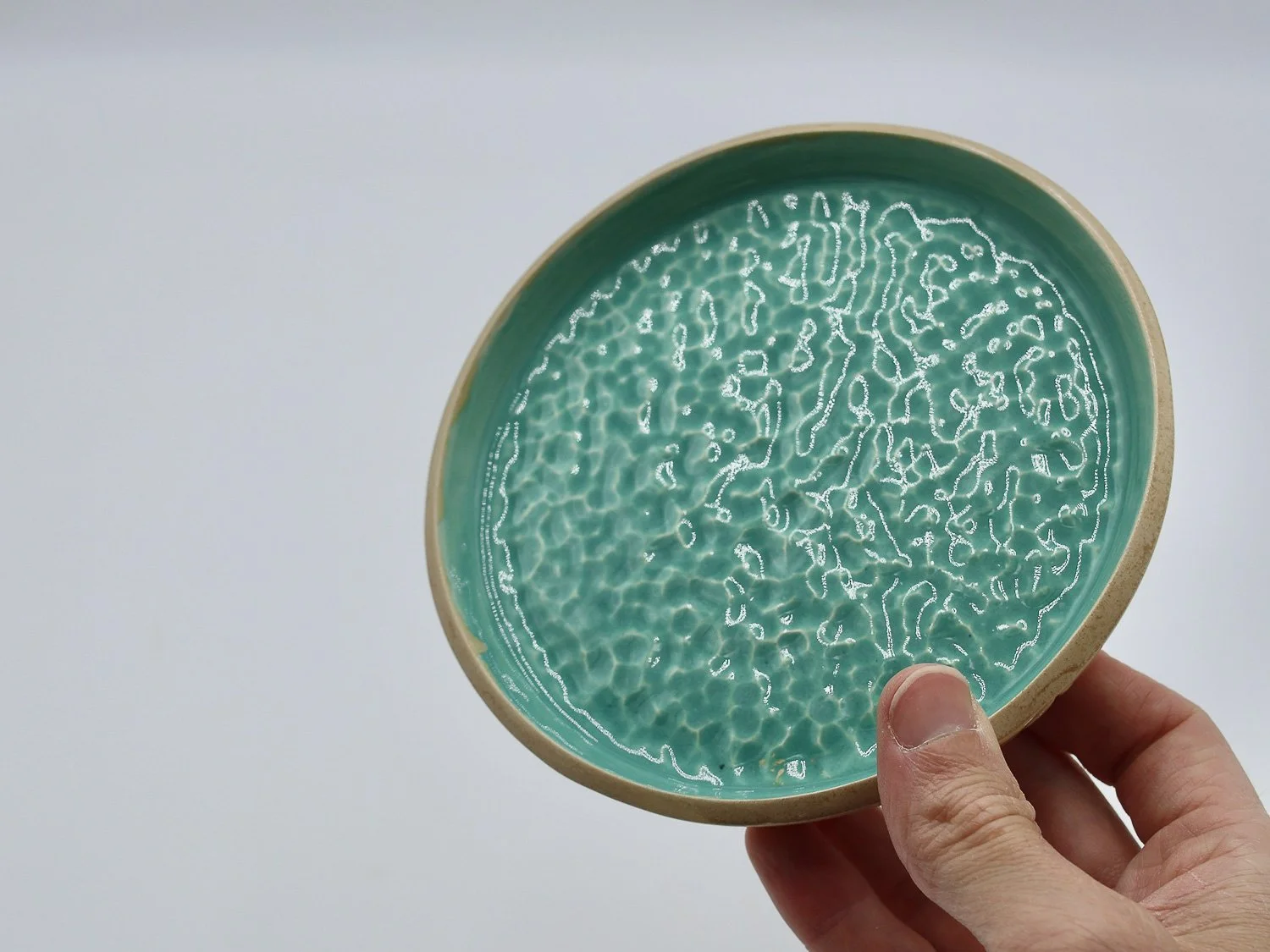 Mão a segurar em prato turquesa e beige em cerâmica, com textura que transmite os reflexos da água
