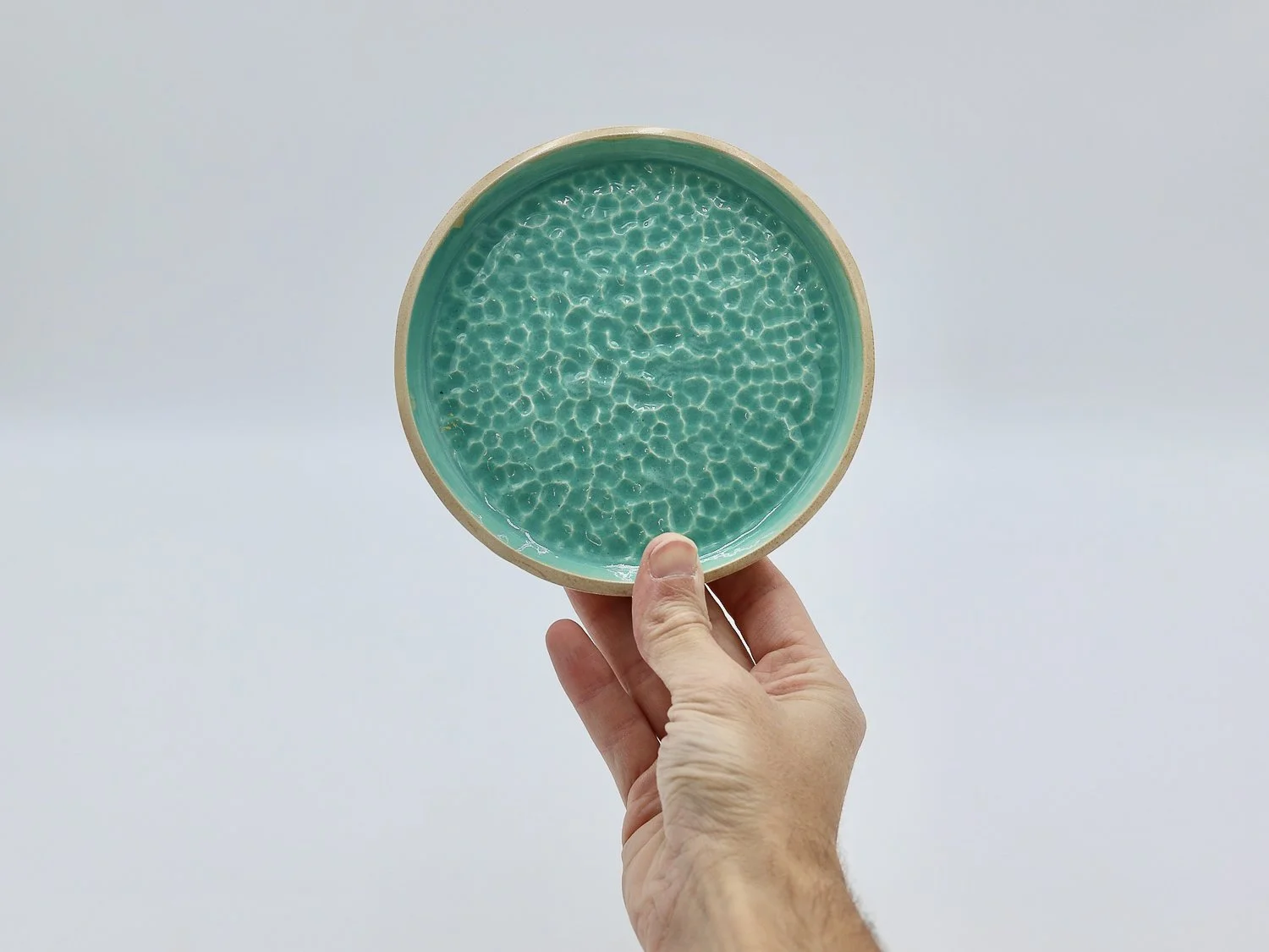 Mão a segurar em prato turquesa e beige em cerâmica, com textura que transmite os reflexos da água