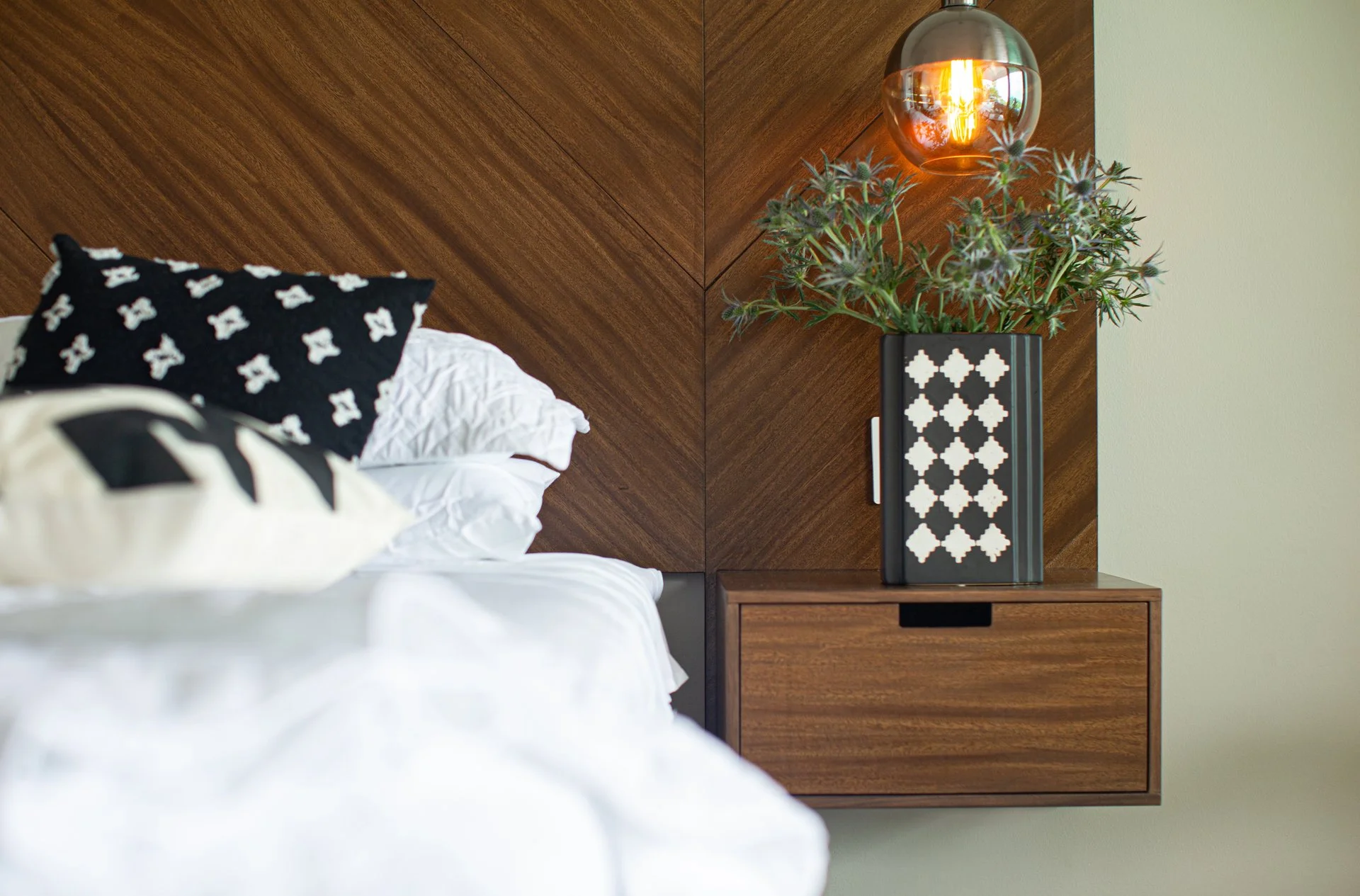 Close-up of a modern bedroom nightstand with a decorative plant in a black and white patterned planter and a hanging light fixture, with a wooden headboard and an unmade bed with black and white pillows in the background.