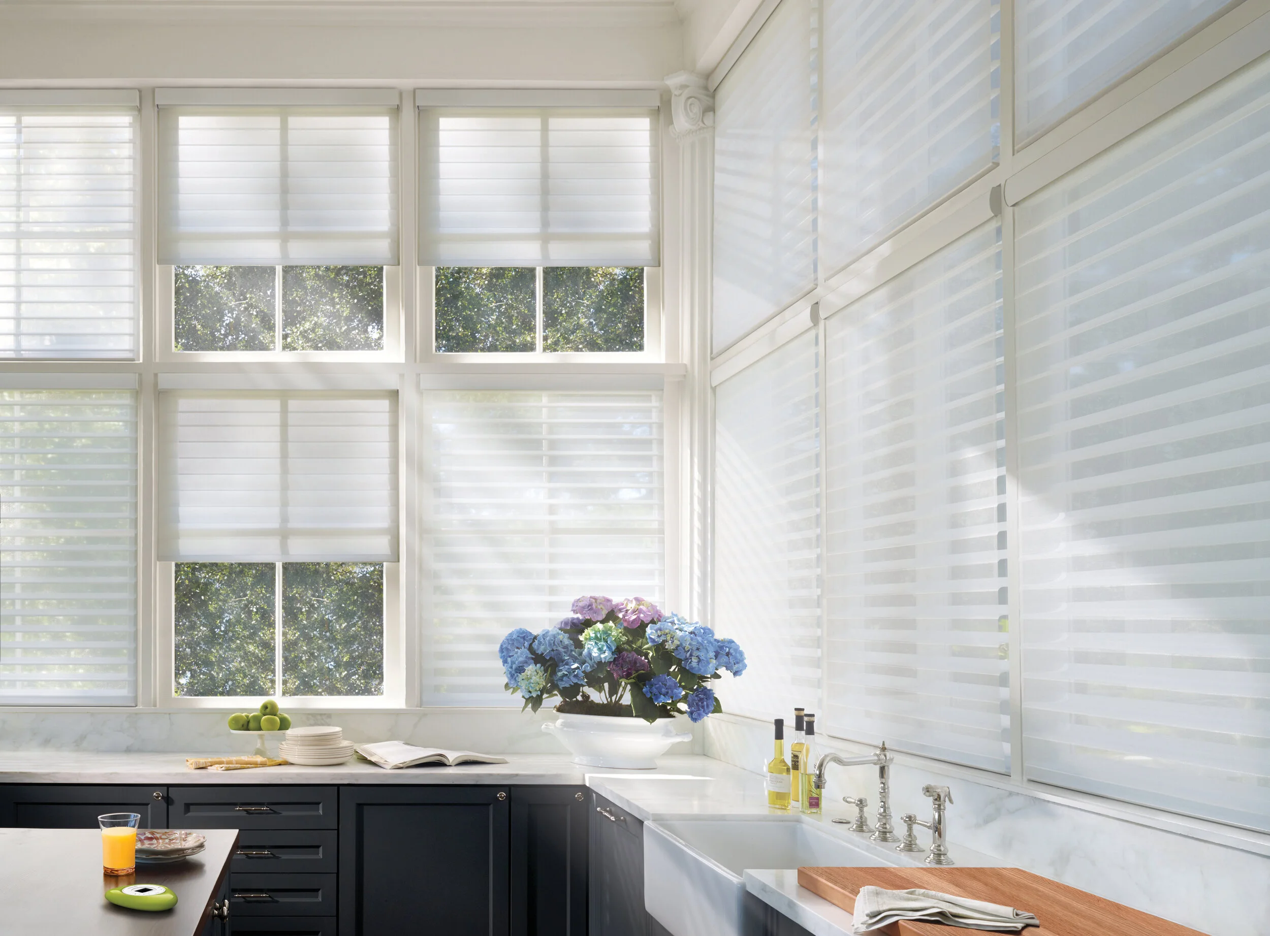 Bright kitchen with large windows covered by white blinds, black cabinets, a white marble countertop, a vase of blue and purple hydrangeas, a bowl of green apples, dishes, and a cutting board.