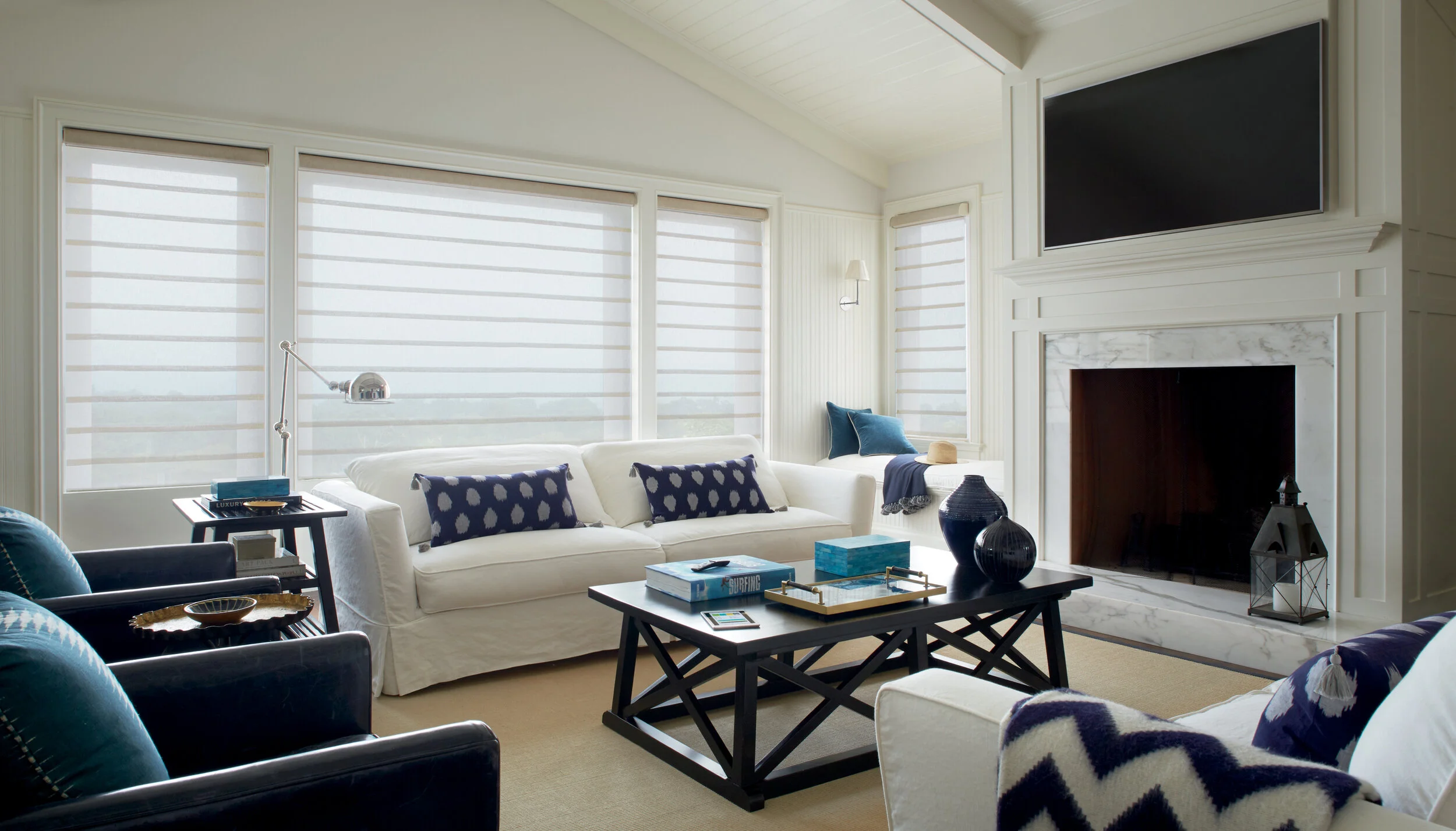 Bright living room with white sofa, navy and white pillows, black and white armchairs, a black coffee table with books and decorative vases, large windows with white blinds, and a fireplace with a mounted TV above.