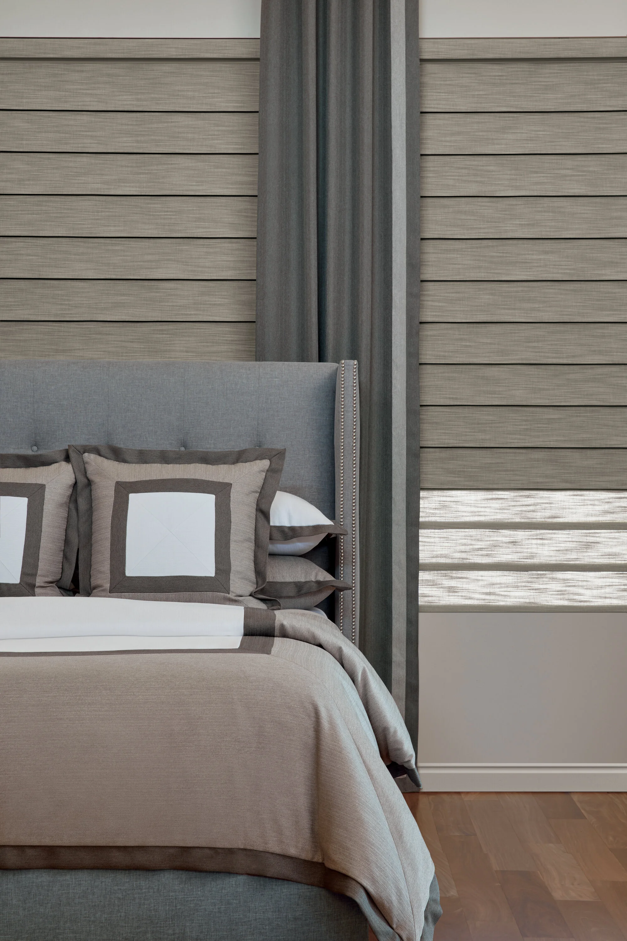 A neatly made bed with gray upholstered headboard and decorative pillows, situated against a wall with horizontal beige wood panels and window with gray horizontal blinds.