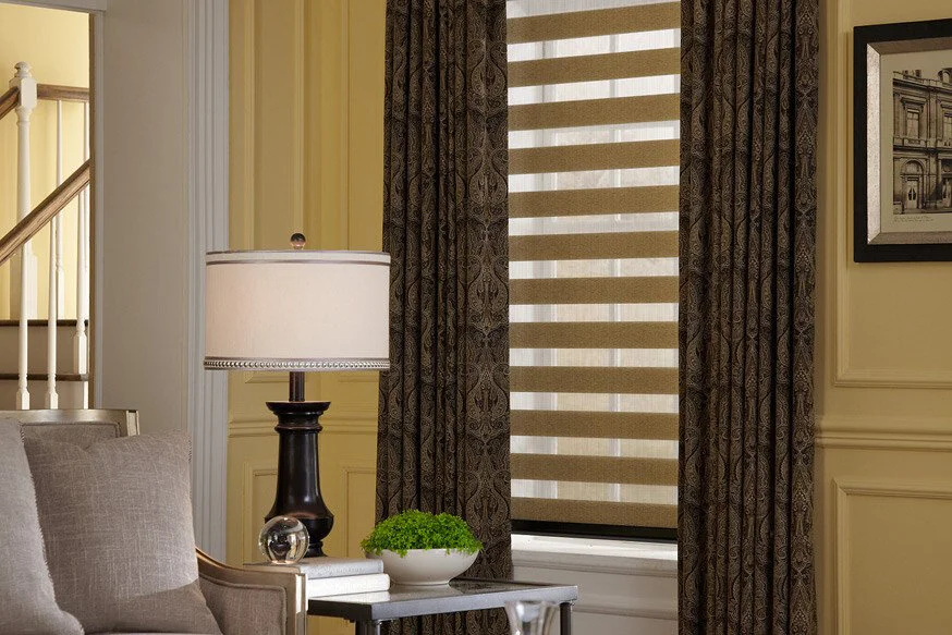 Close-up of a corner in a living room with a window, dark patterned curtains, a table lamp, a framed photo, and a potted plant.