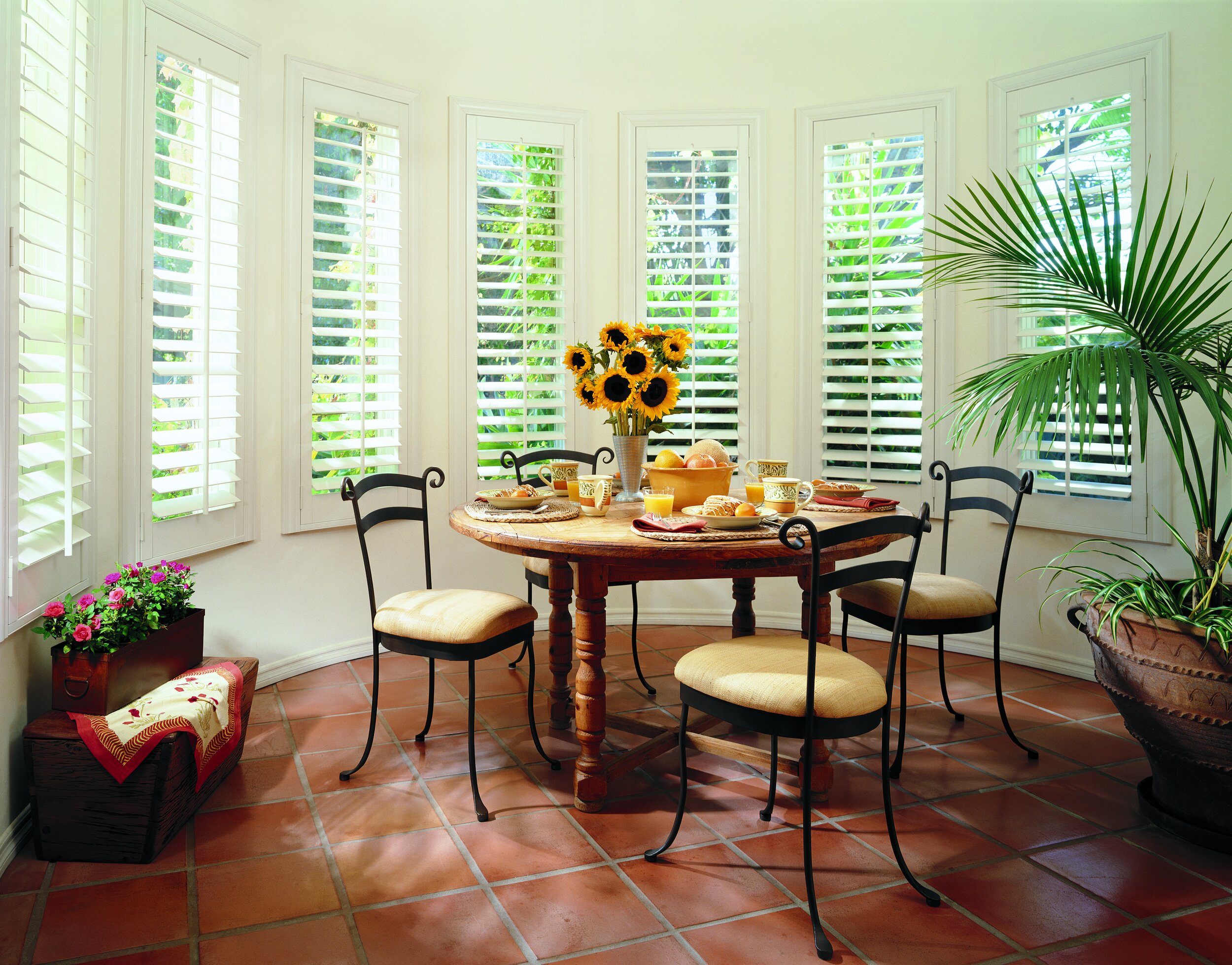 A sunlit dining area with a round wooden table set for breakfast, a vase of sunflowers at the center, and surrounded by four chairs, near large windows with white plantation shutters, terracotta tiled floor, and houseplants in the corners.