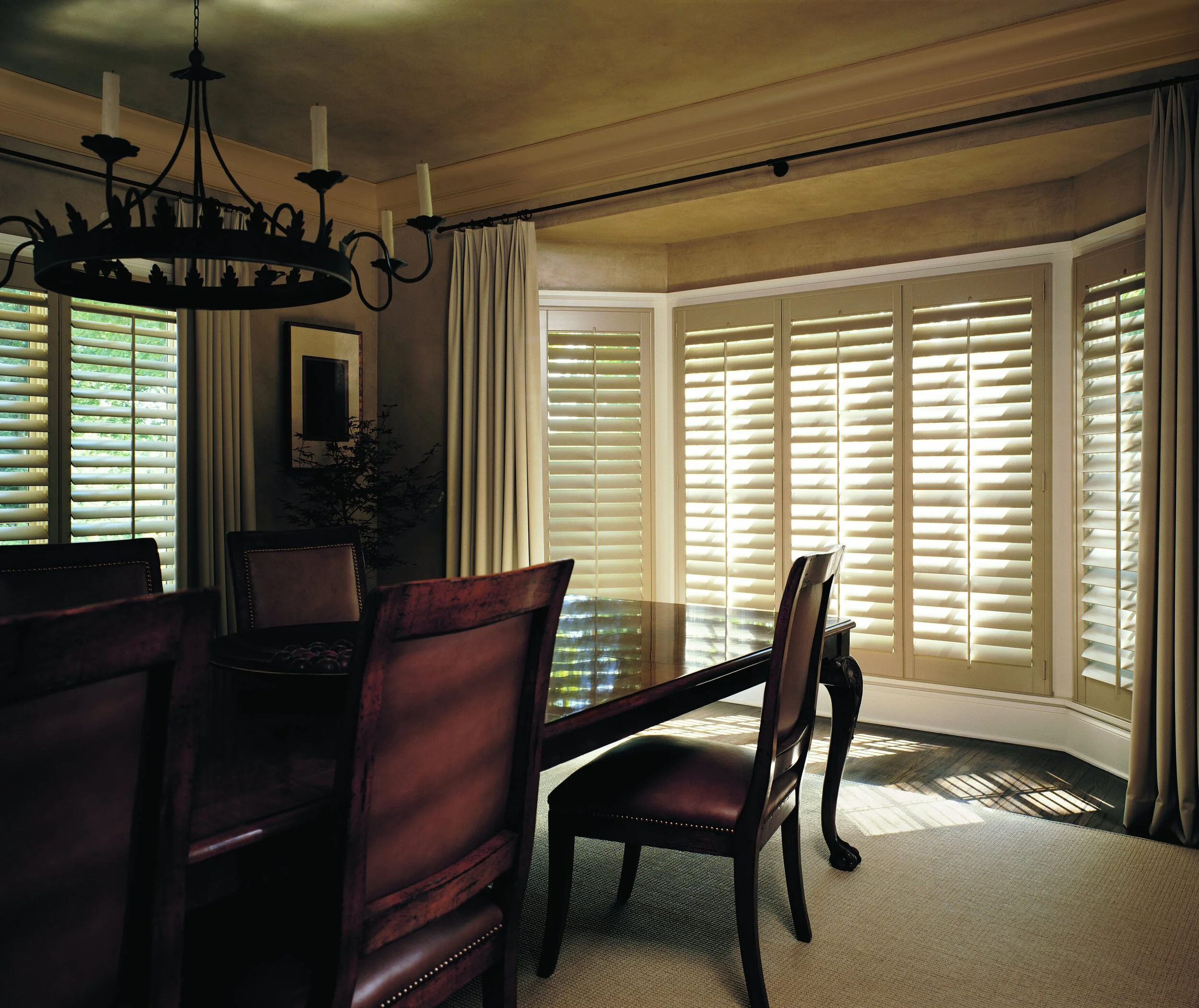A dining room with a wooden table, dark upholstered chairs, a chandelier, and large windows with white plantation shutters and cream curtains allowing sunlight in.