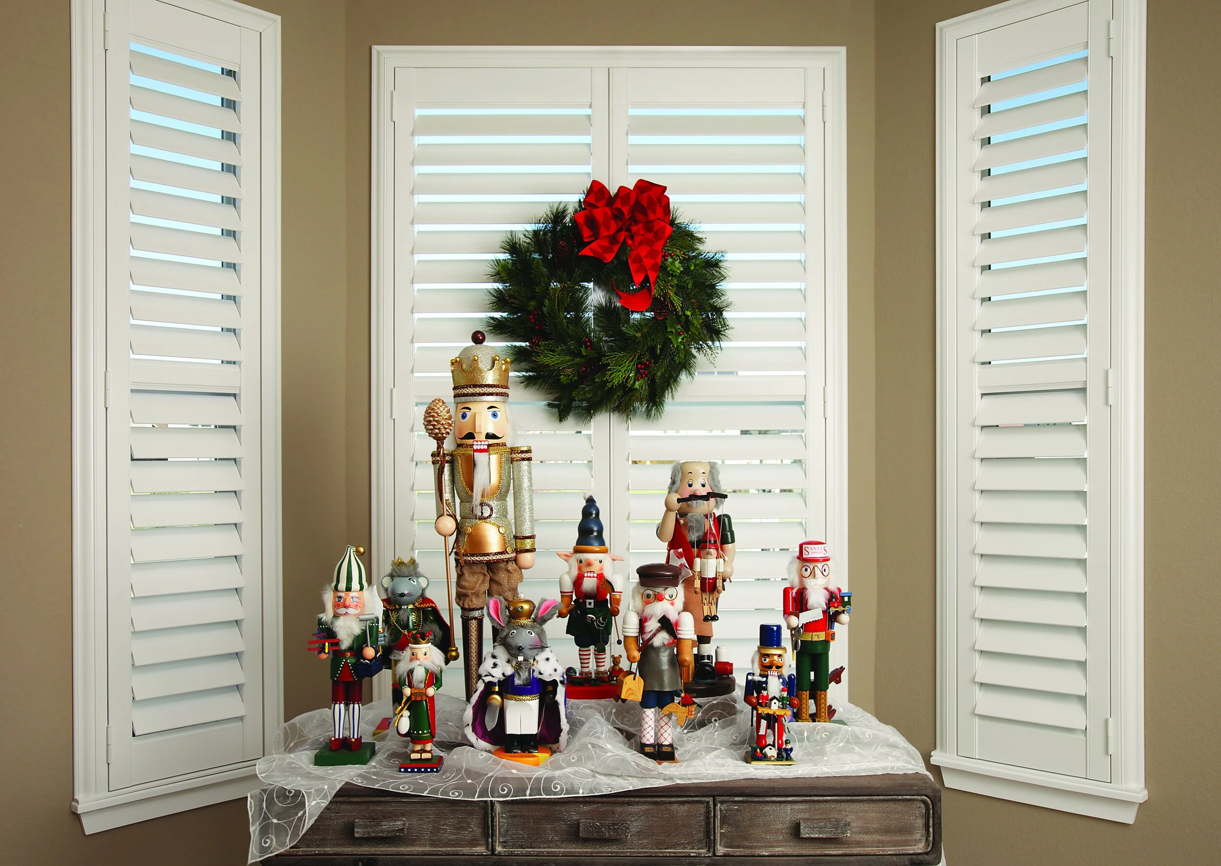 Collection of nutcracker and Santa Claus holiday decorations on a wooden table, with a green wreath with a red bow hanging on white window shutters in the background.