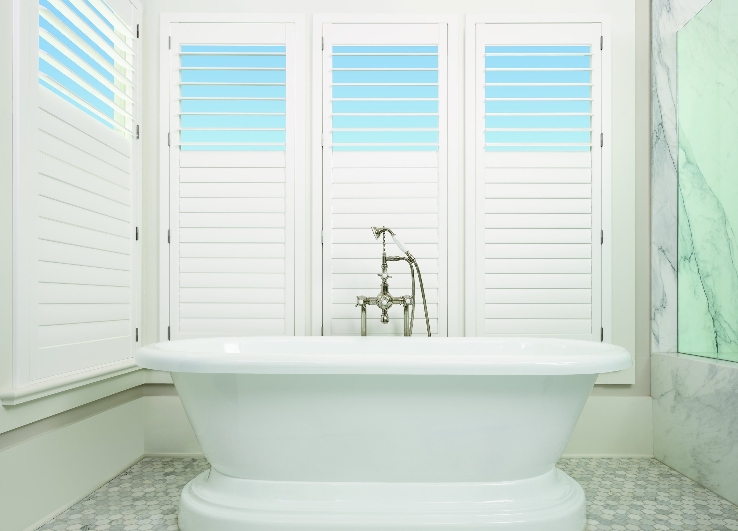 White bathtub with vintage faucet in a bright, modern bathroom with white shutters and marble accents.