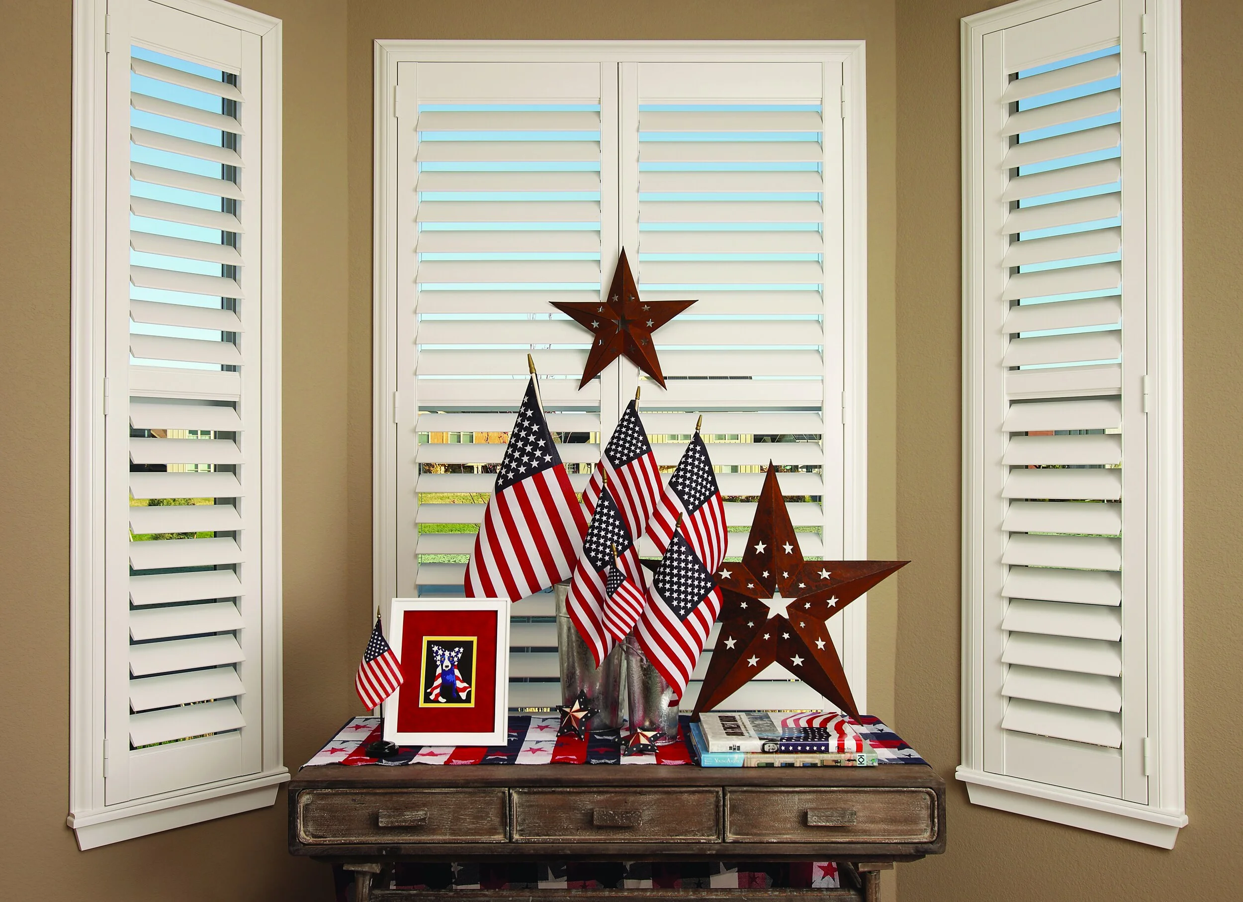 Decorative patriotic display with American flags, star ornaments, and a framed picture on a wooden table in front of a window with white shutters