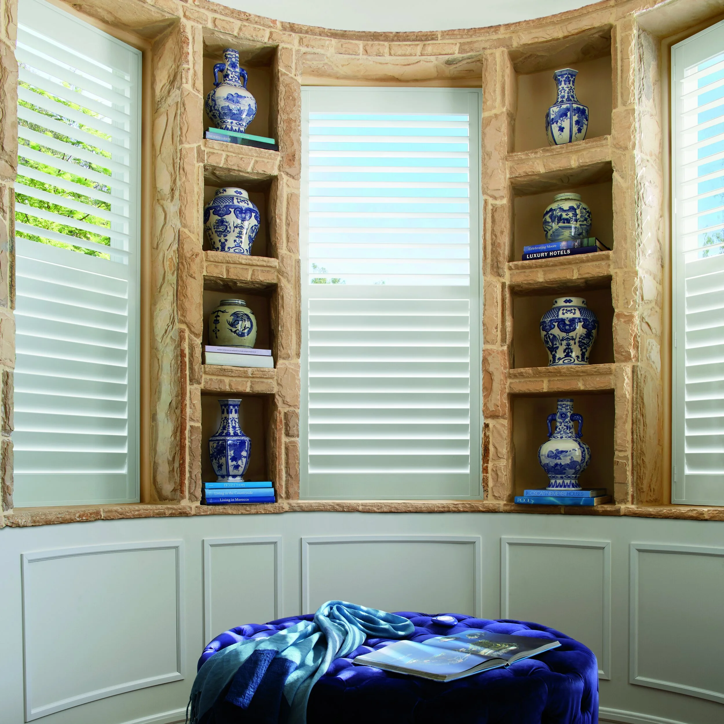 A window nook with built-in shelves showcasing blue and white Chinese porcelain vases, brightened by sunlight coming through shutters, with a blue velvet ottoman in front with an open book and a scarf on top.
