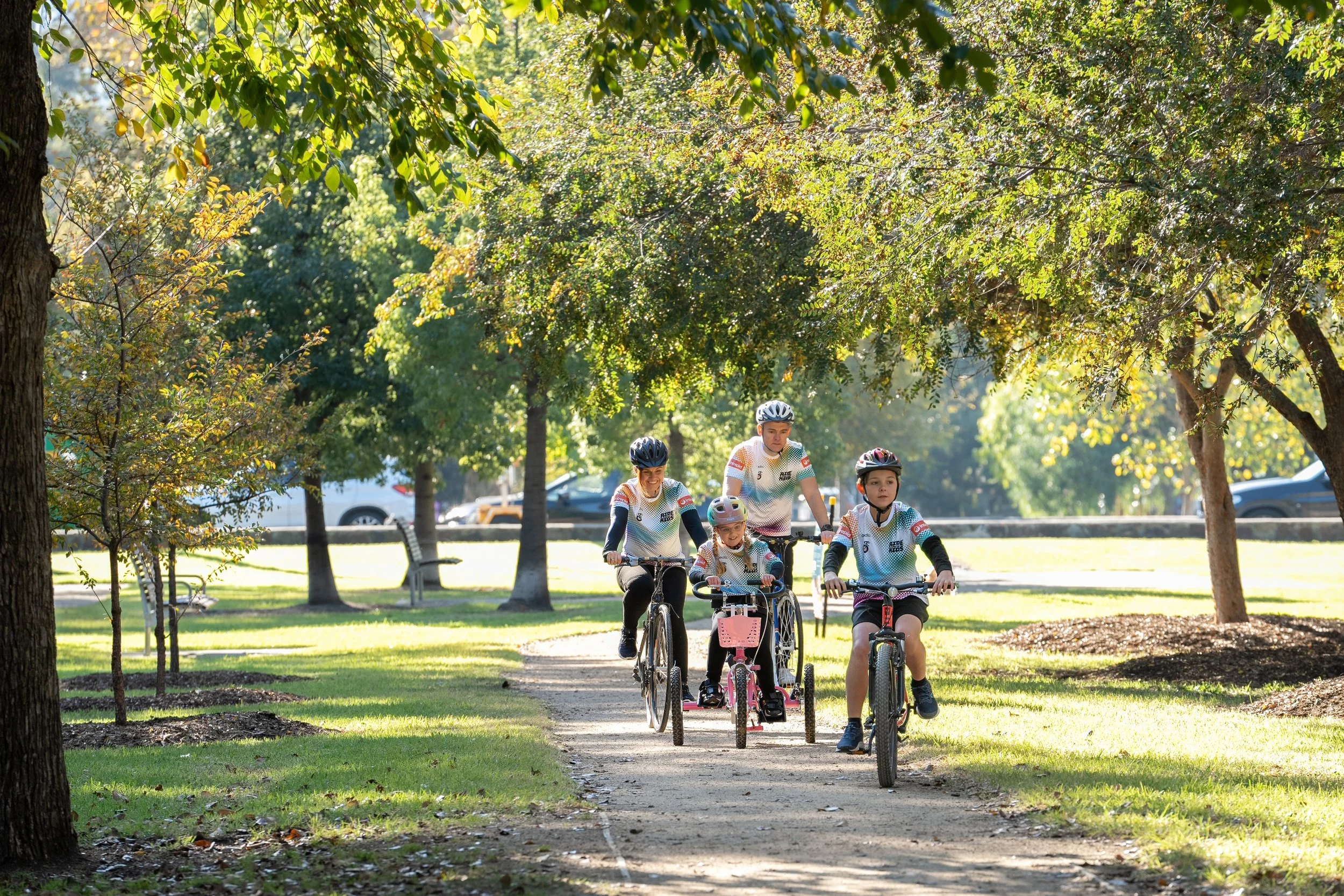 Children riding bikes on a park path surrounded by trees, with sunlight filtering through the leaves.