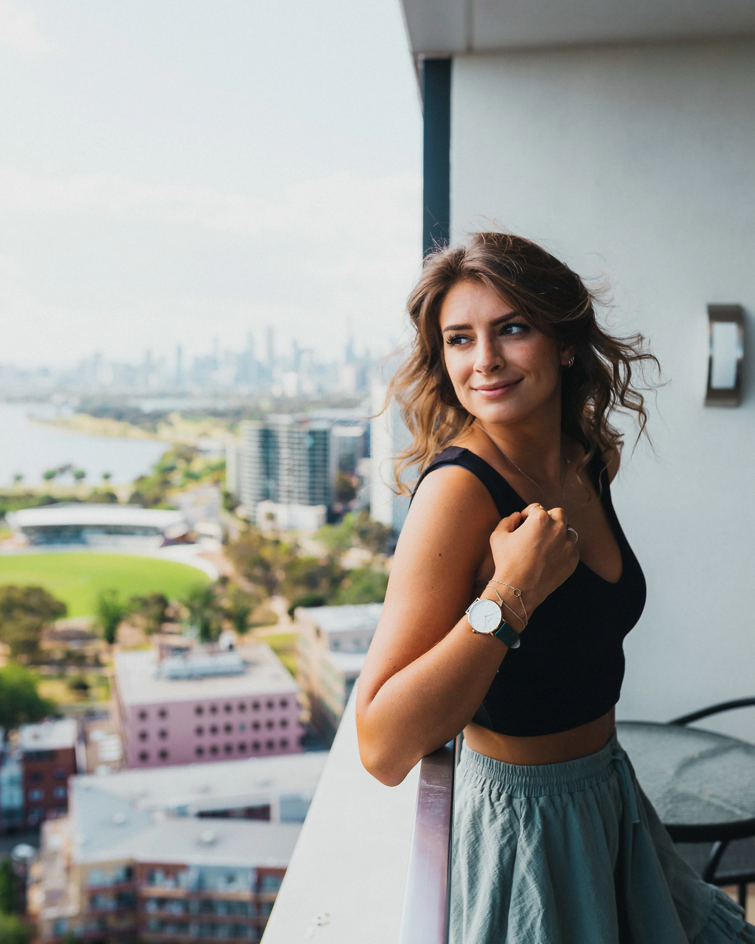 A young woman with wavy brown hair smiling and looking to the side, standing on a balcony with a cityscape in the background, wearing a black top, beige skirt, and a watch on her left wrist.