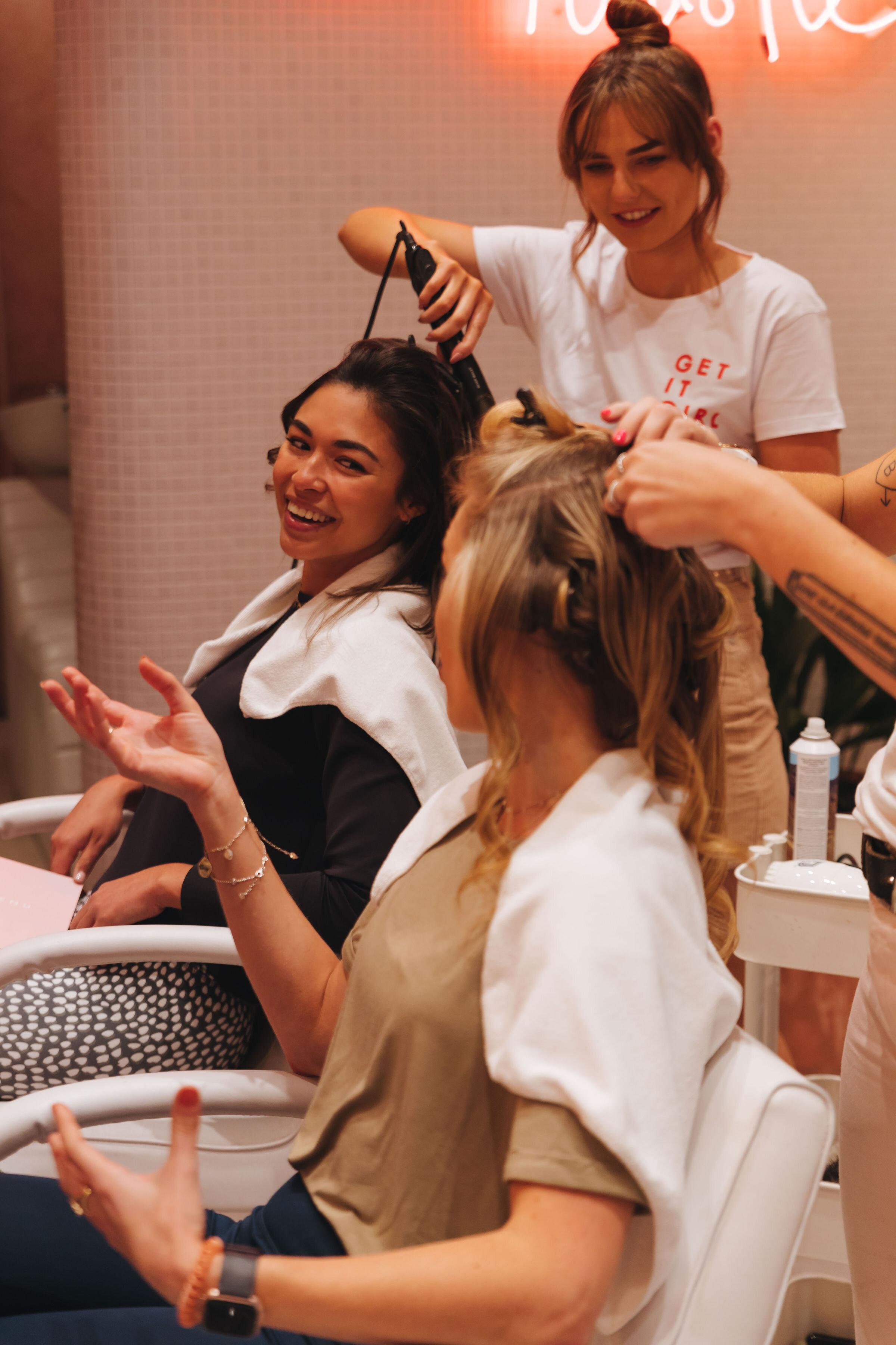 Two women sitting at a table with beauty tools, smiling and talking while one styles the other’s hair in a salon or spa setting.