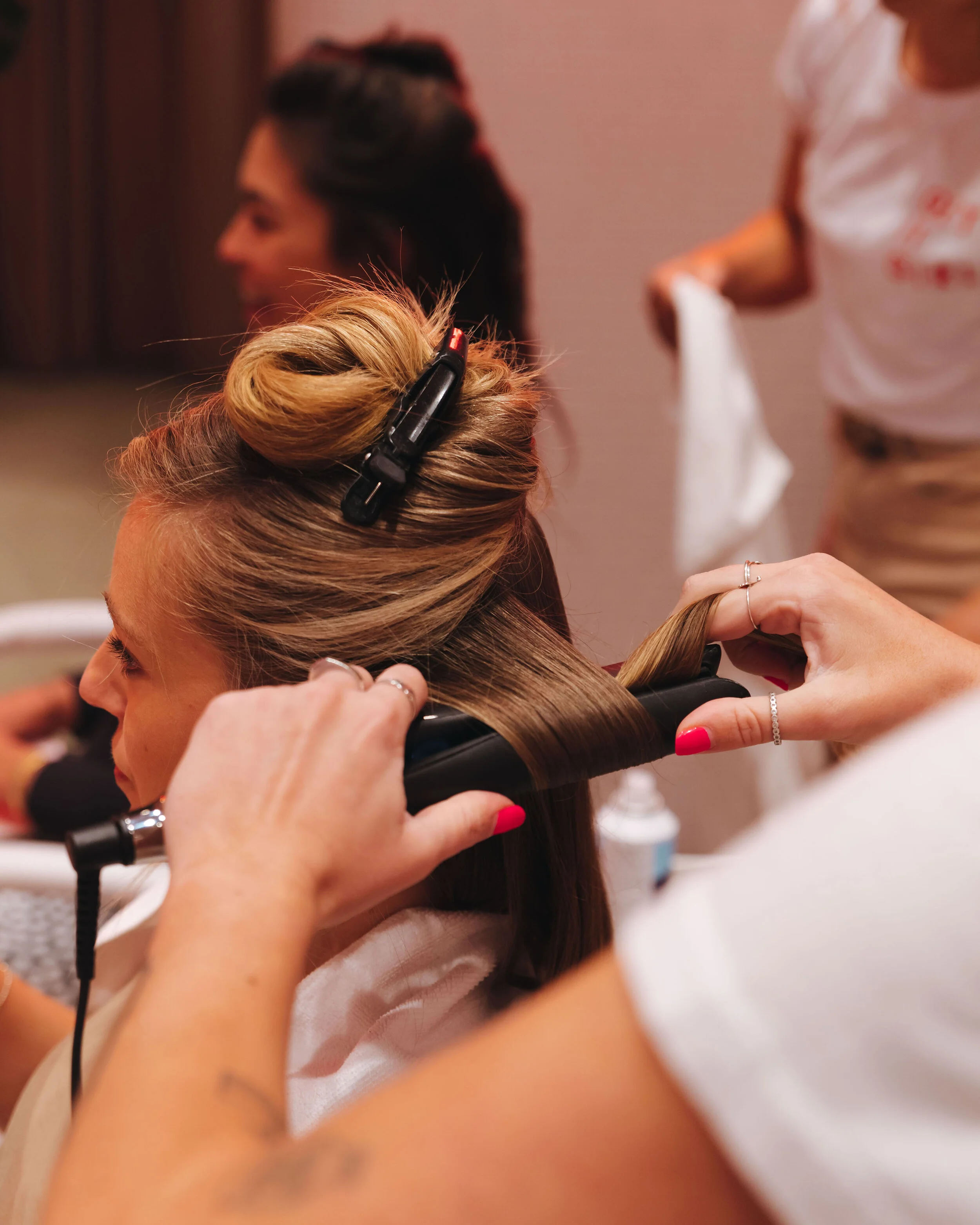 A woman getting her hair styled in a salon, with a stylist working on her hair. The woman has blonde hair in a bun with a hair clip, and the stylist is wearing a white shirt with red writing, holding a section of her hair.