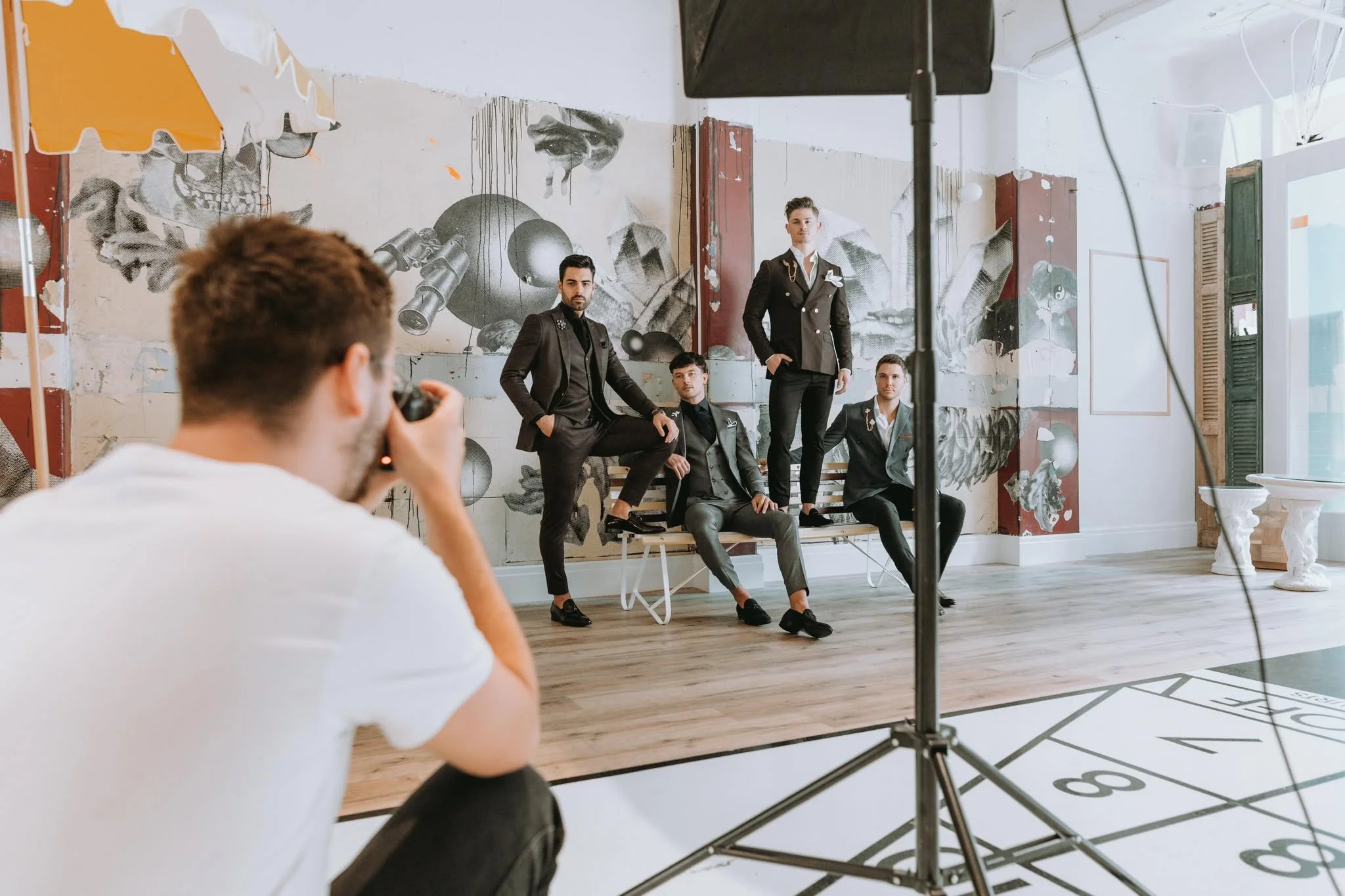 A photoshoot features five male models dressed in black suits with stylized designs, posing in front of a large graffiti-style mural on the wall. The mural includes abstract shapes, eye imagery, and other artistic elements. A photographer in a white shirt is taking their picture, with a studio light and other photography equipment visible in the room, which has a bright, artistic atmosphere.