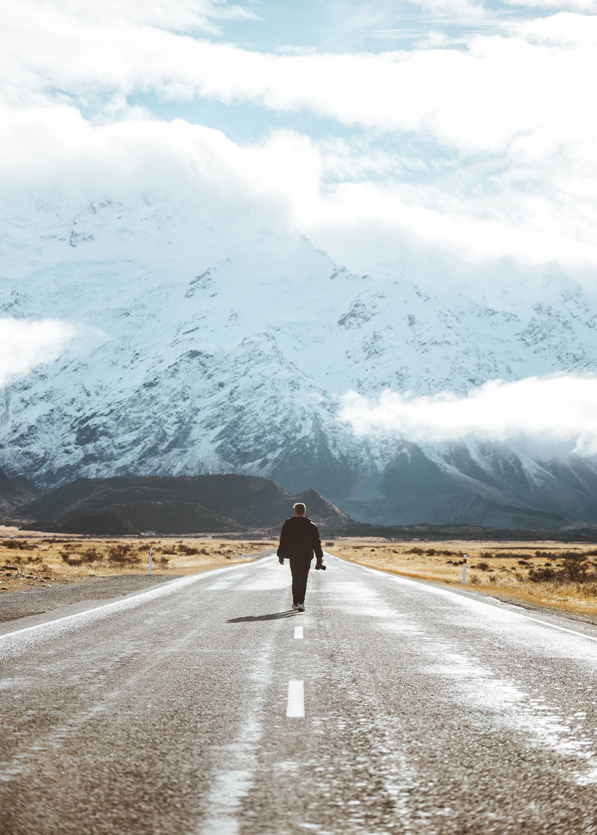 A person with a camera walking down an empty road towards mountains with snow and clouds.