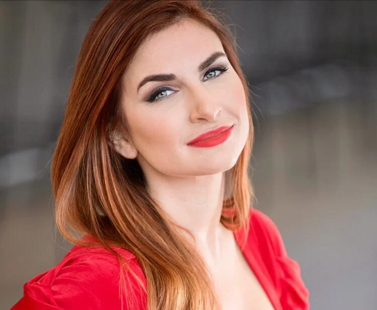 Close-up of a woman with red hair, fair skin, and red lipstick, smiling slightly, wearing a red top.