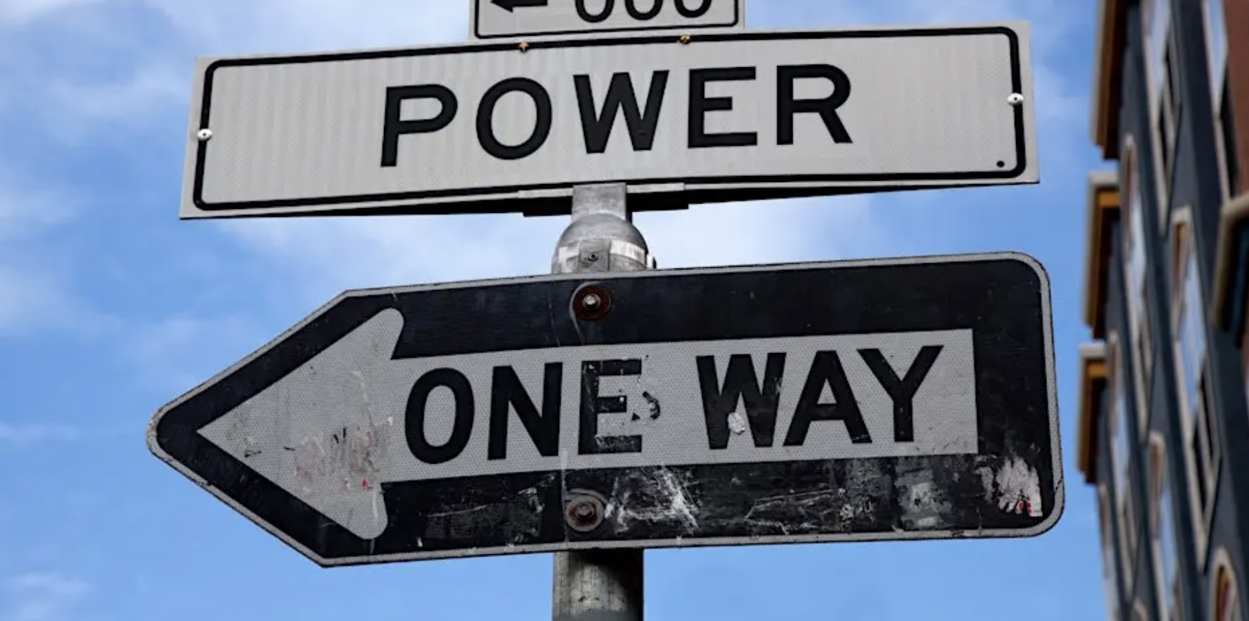 Street signs on a metal pole against a blue sky: a rectangular sign reading “POWER” above a left-pointing arrow sign that says “ONE WAY.” A building facade is partially visible on the right side.