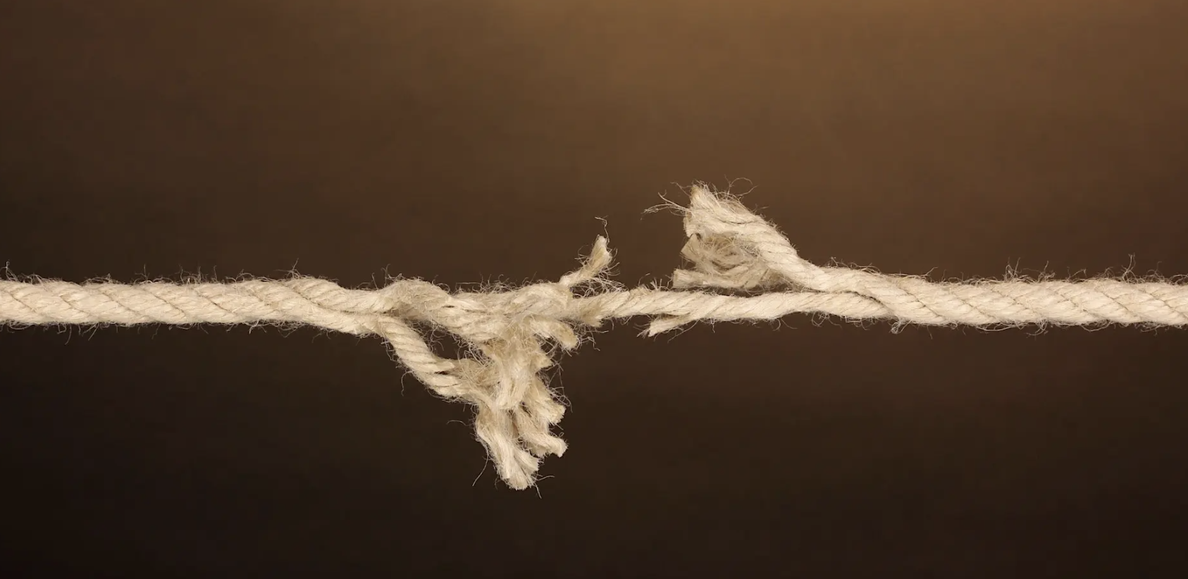 A fraying rope pulled taut from both ends, nearly snapped at its center point, against a dark brown background.