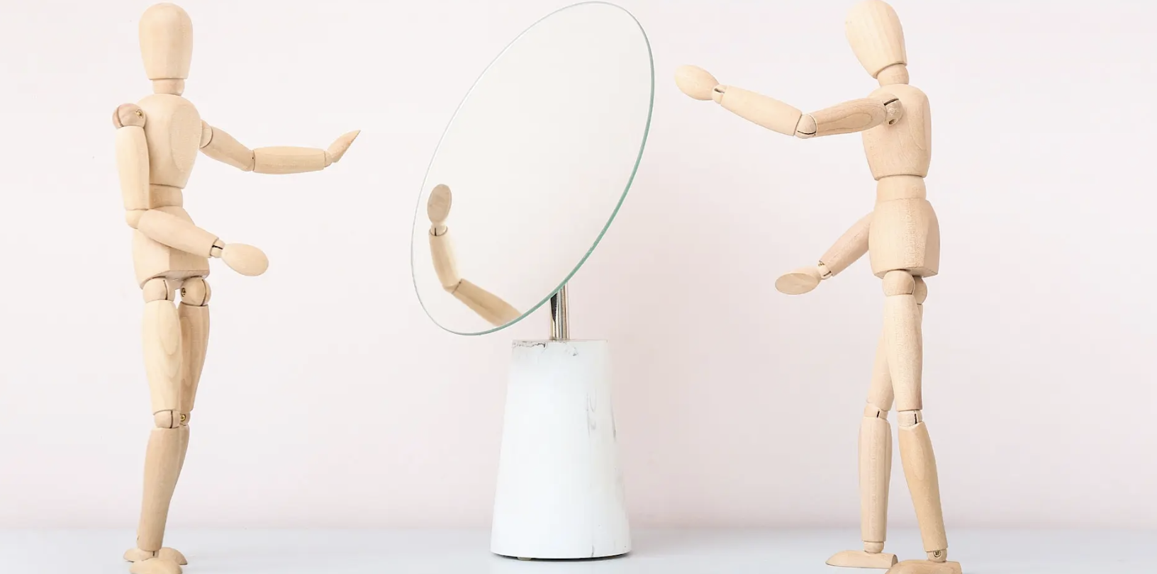 Two wooden artist mannequins stand on either side of a round mirror on a white stone base, each with an arm raised toward the reflection.
